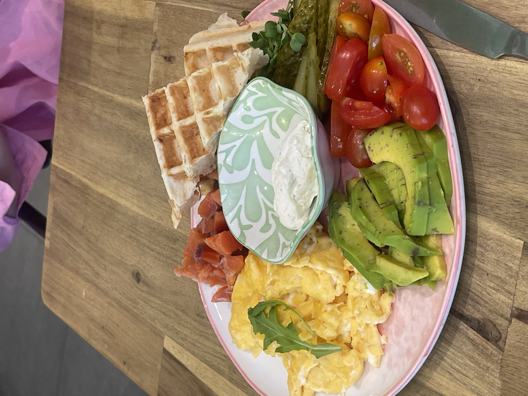 The image displays a vibrant breakfast or brunch platter, presented on a round, light pink and white rimmed plate set on a wooden table. The plate is generously filled with a variety of food items.
In the upper left quadrant, several pieces of golden-brown waffles, cut into rectangles, are arranged. Below them, a portion of smoked salmon with an orange hue is visible. A small, decorative bowl, white with a distinctive green leaf-like pattern, sits centrally on the plate, containing a creamy white dip or sauce, possibly sour cream or cream cheese.
To the right of the dip bowl and extending to the top right, several long, green pickled cucumbers or gherkins are neatly stacked. Adjacent to these, a cluster of bright red cherry tomatoes, mostly halved, adds a fresh and colorful element. Below the tomatoes and along the right edge of the plate, perfectly fanned slices of ripe green avocado are arranged.
At the bottom center of the plate, a fluffy mound of scrambled eggs is served, garnished with a fresh green arugula leaf. A few microgreens are also scattered near the waffles and pickles. The wooden table surface, with its visible grain and slight imperfections, serves as the background. A silver knife is partially visible on the upper right side of the table, and a folded piece of purple fabric is seen along the far left edge of the frame. The scene is well-lit, suggesting daytime. No people are visible, and there is no discernible text in the image.