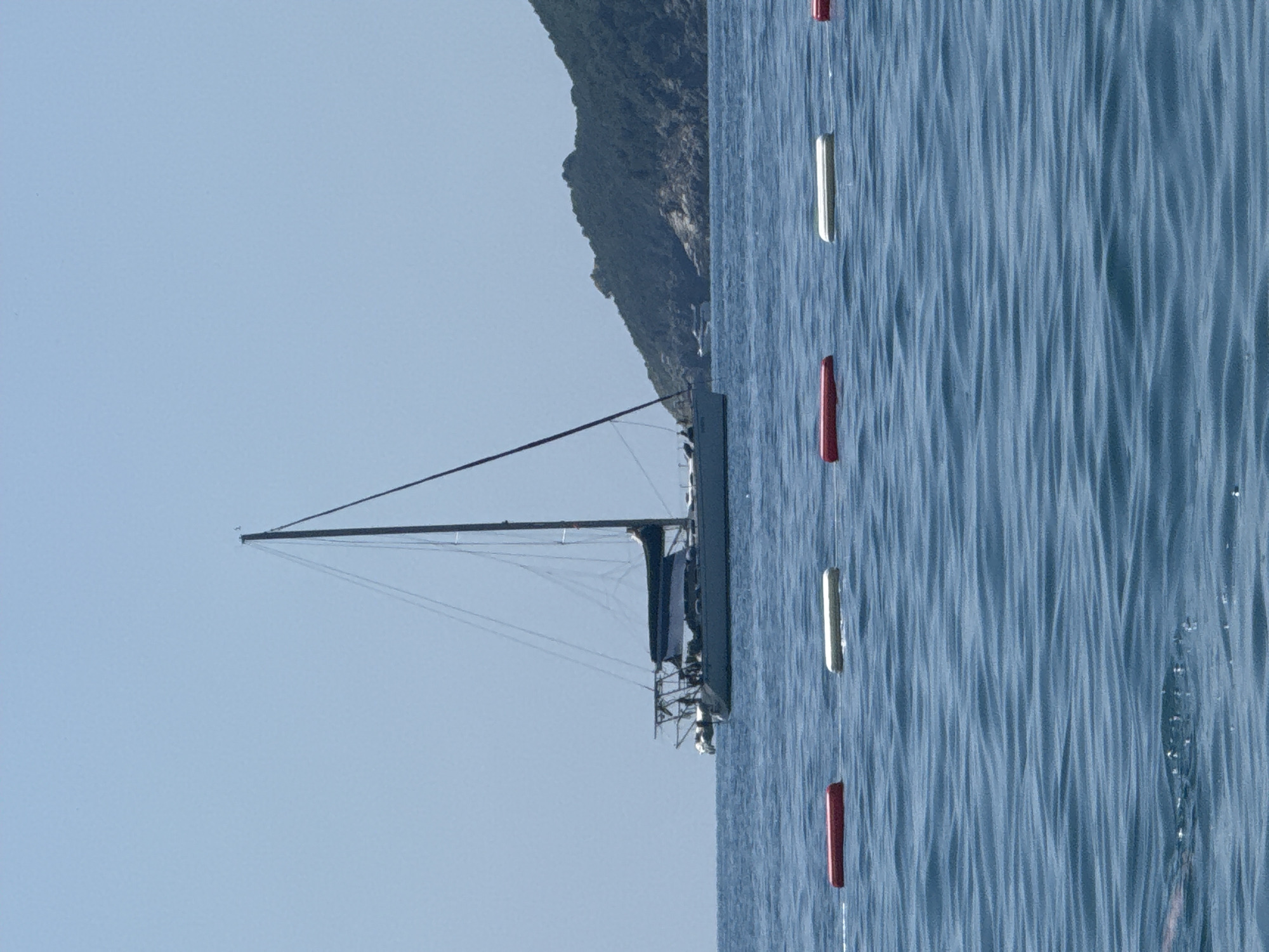 The image depicts a tranquil scene on the water, likely in or near Boreti, Montenegro. A white sailboat with dark sails is anchored in the blue water. The sailboat has a tall mast and appears to be of considerable size. In the background, a lush green hill rises from the sea, dotted with rocky outcrops. In the foreground, several red and white buoys are spaced out in a line across the water, possibly marking a swimming area or a boundary. The sky is clear and blue, suggesting a sunny day. The water is calm, with gentle ripples reflecting the sky. No people are clearly visible in the water.
