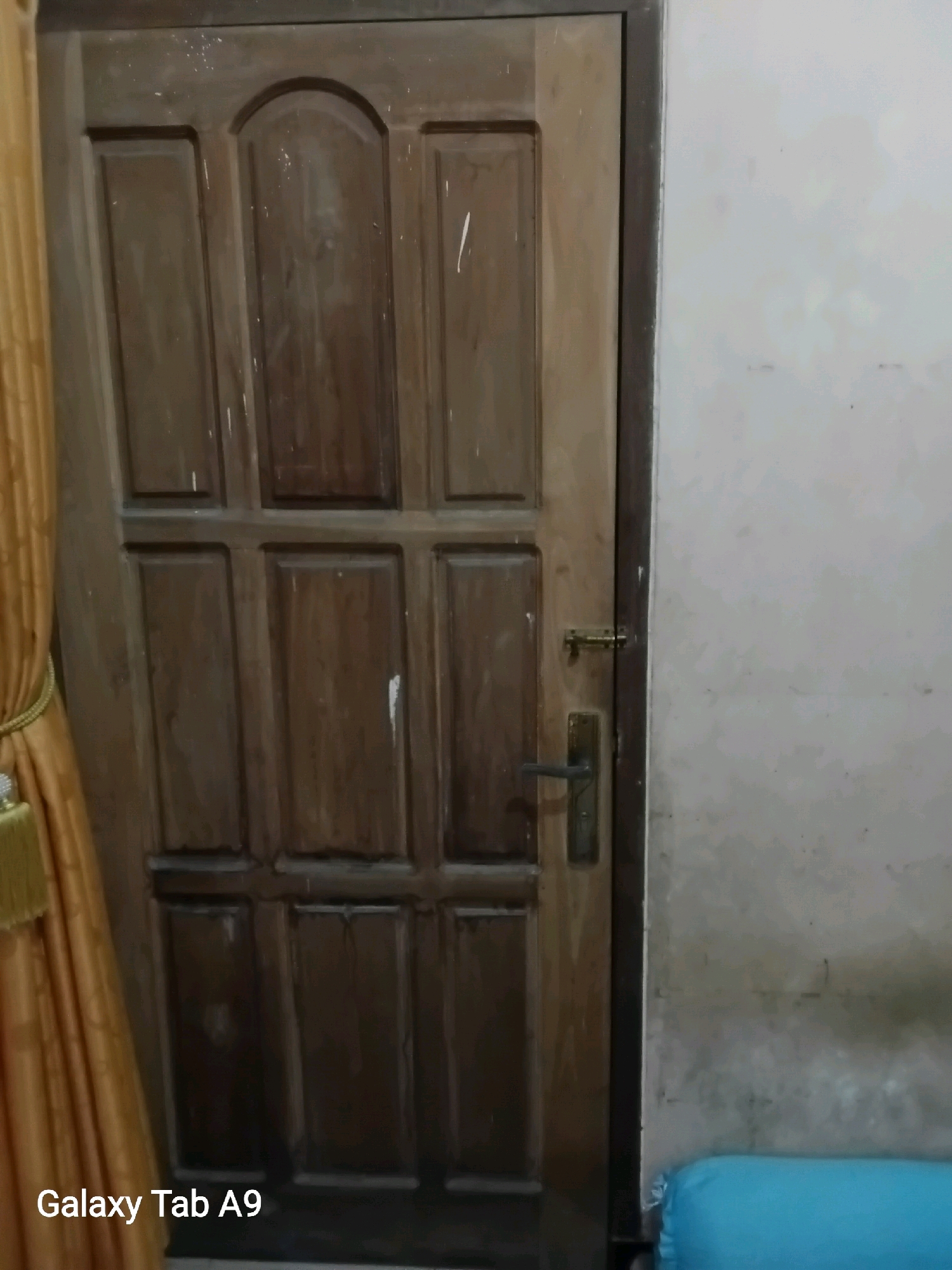The image shows a wooden door with several panels, a brass doorknob, and a latch. The door is partially covered by a golden curtain on the left side. To the right of the door, a light-colored wall is visible, with a blue object, possibly a cushion, resting on the floor. The overall scene appears to be indoors, likely a room. The image is a photograph taken by a Samsung Galaxy Tab A9, as indicated by the watermark at the bottom left corner. There are no people or specific locations in Trenggalek, Indonesia, depicted in the image.