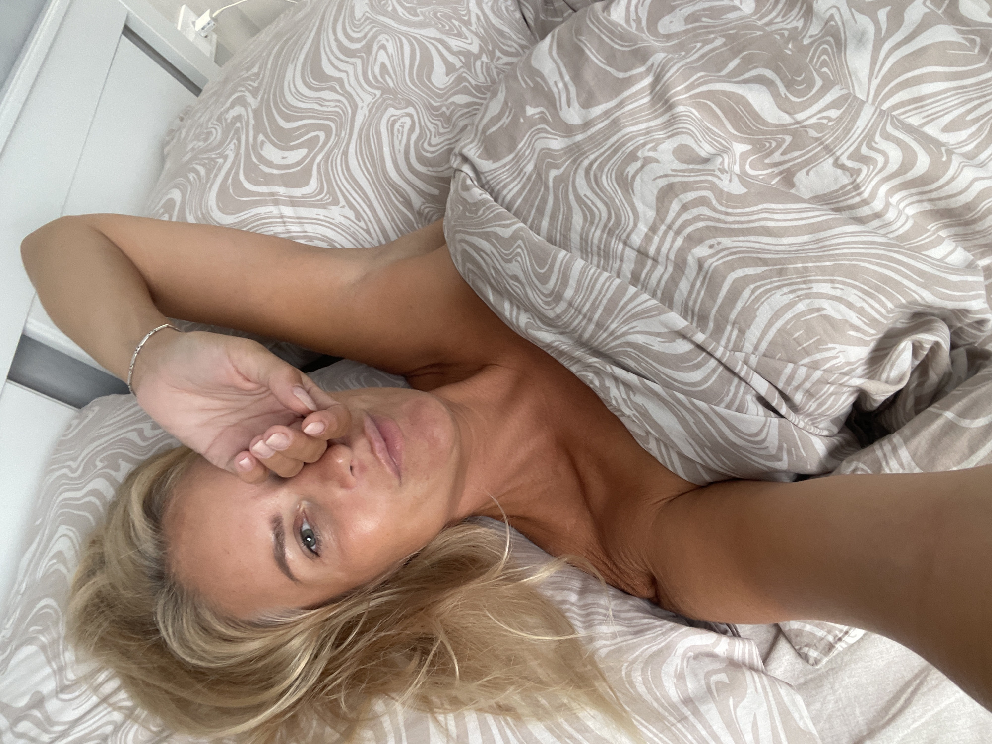 A blonde woman is lying in a bed, partially covered by a duvet with a swirling pattern. She is looking at the camera with her right hand covering her eye. Her left arm is resting above her head, and she is wearing a silver bracelet. The bed is positioned against a white headboard, and a power outlet is visible on the wall. The lighting suggests it is daytime. The scene is indoors, likely a bedroom. There is no information in the image that can definitively place this location in Novorossiysk, Russia.