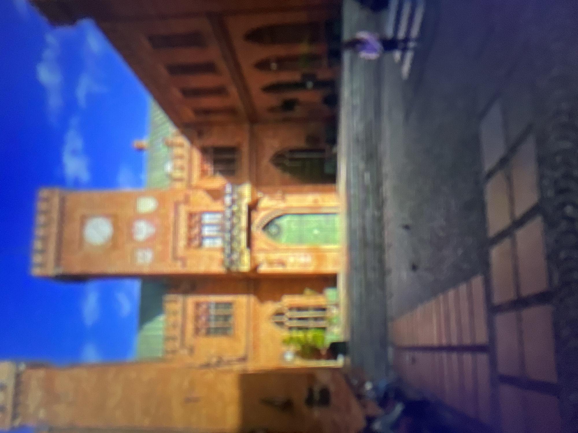 The image depicts a stylized, animated scene that appears to be from a video game or computer-generated animation. The primary subject is a large, ornate building with a prominent tower, constructed from warm-toned brickwork. The building features multiple windows, some of which are arched, and decorative elements. A large, light-colored clock face is visible on the tower. A bright green door or window is also a noticeable feature of the building.
In the foreground, a cobblestone path or courtyard is visible, with shadows cast upon it, suggesting it is daytime. The sky is a vibrant blue with fluffy white clouds, indicating fair weather. A single figure, appearing to be a person in purple clothing, is walking away from the camera on the cobblestone path. The overall atmosphere of the scene is serene and somewhat whimsical.
The scene is oriented vertically in the image, with the sky and upper parts of the building appearing at the bottom and the cobblestone path and lower parts of the building at the top. This unusual orientation may be a result of how the image was captured or processed.
The location context provided is Kołobrzeg, Poland, though there are no explicit visual cues within the image itself that directly confirm this location, such as identifiable landmarks or text. The architecture suggests a European setting, possibly with historical influences.