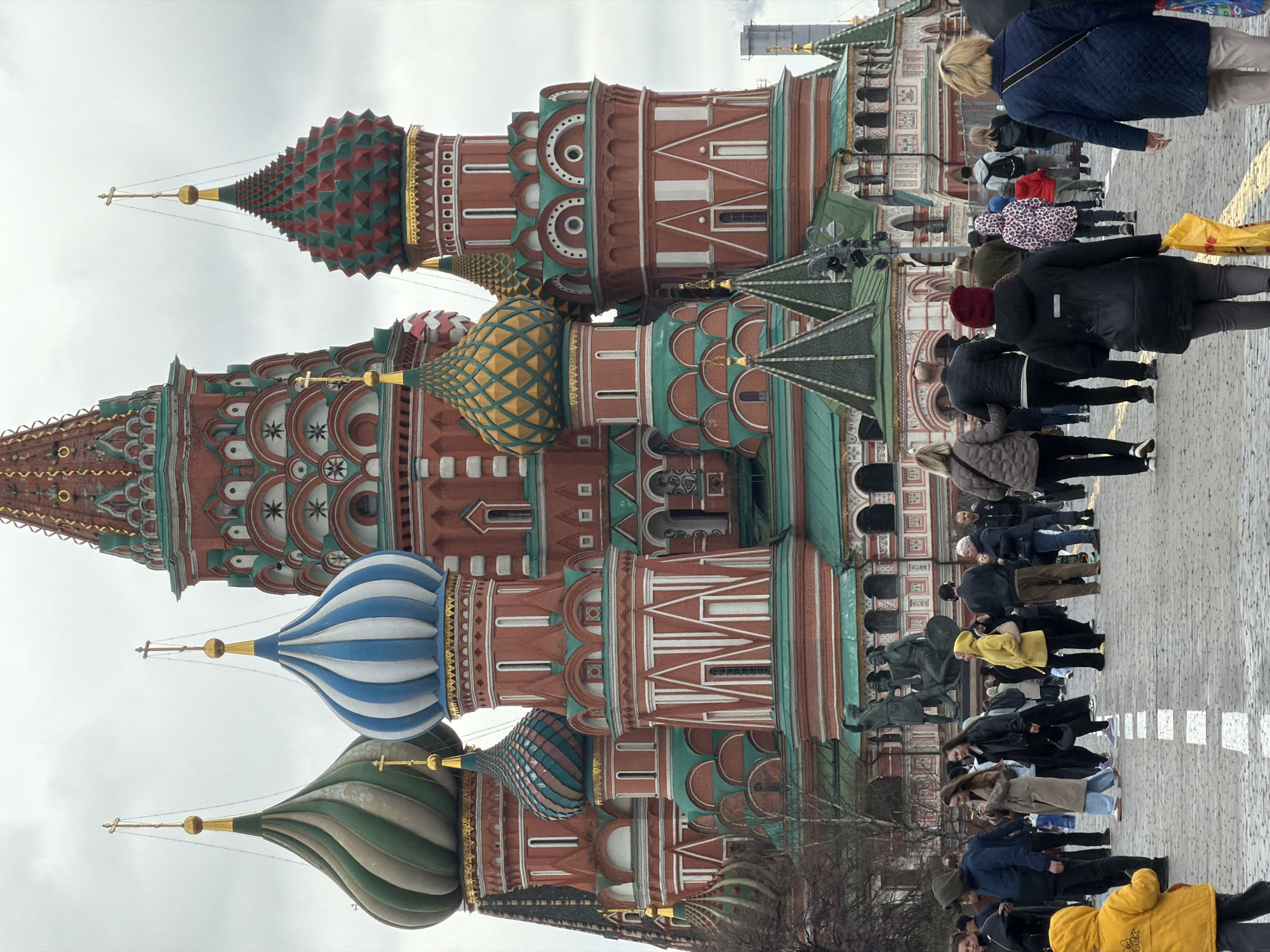 The image captures the vibrant architecture of St. Basil's Cathedral in Moscow, Russia. The cathedral is characterized by its distinctive onion-shaped domes, each decorated with unique and colorful patterns. The domes are mounted on structures with a red and white color scheme, further adorned with green and gold accents.
In the foreground, a crowd of pedestrians walks along a cobblestone path. People of various ages and wearing diverse clothing styles are present.
The sky is overcast, casting a muted light over the scene. The composition of the image emphasizes the height and grandeur of the cathedral, contrasting with the human scale of the pedestrians in the foreground.