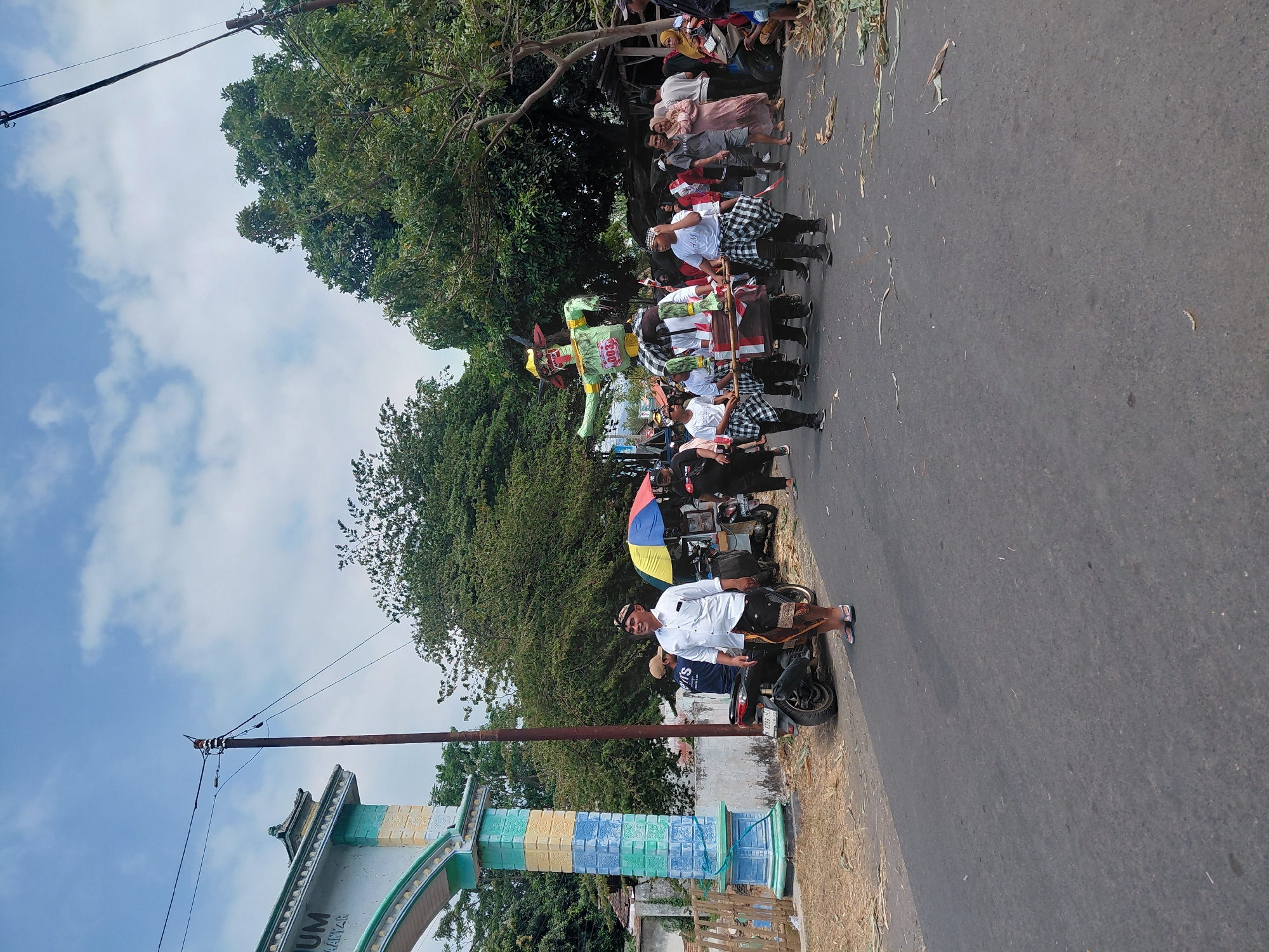 In the city of Bimo, Indonesia, a procession moves along an asphalt road under a partly cloudy sky. A man in a white shirt and traditional dark patterned sarong stands near an ornate archway adorned with blue and yellow pastel colors, bearing the text "UM" and "AANYAP" in black. To his right, a motorcycle is parked. Further down the road, a group of people are carrying a large, brightly colored effigy, likely a "ogoh-ogoh" figure, green and adorned with yellow and red accents, over their shoulders using wooden poles. The effigy has a large head with outstretched arms and a red number tag. Behind the effigy carriers, more people walk, some dressed in traditional checkered patterns. A small stall with a colorful umbrella is visible, and foliage lines the side of the road, with some debris scattered on the asphalt. The scene suggests a cultural celebration or parade in progress.