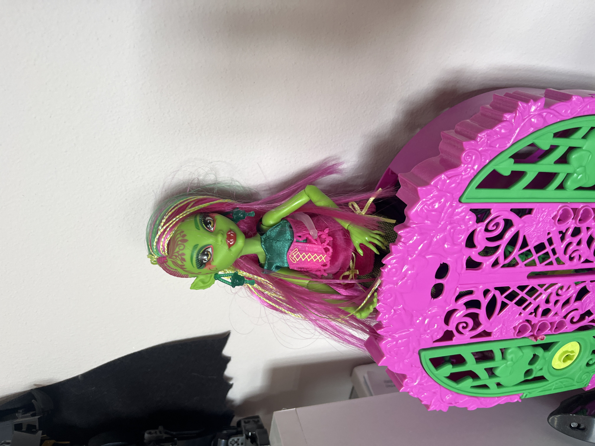 A close-up, eye-level shot shows a Monster High doll with green skin, pink and green braided hair, and large pointed ears, wearing a green and pink corset top and a pink skirt. The doll is partially enclosed within a bright pink, ornate toy accessory that resembles a miniature decorative carriage or greenhouse with green latticework doors. The doll is positioned against a plain white wall, with a dark object, possibly a cape or fabric, visible in the upper left corner of the frame. There is no information available to determine the location in the city of Onokivtsi, Ukraine.
