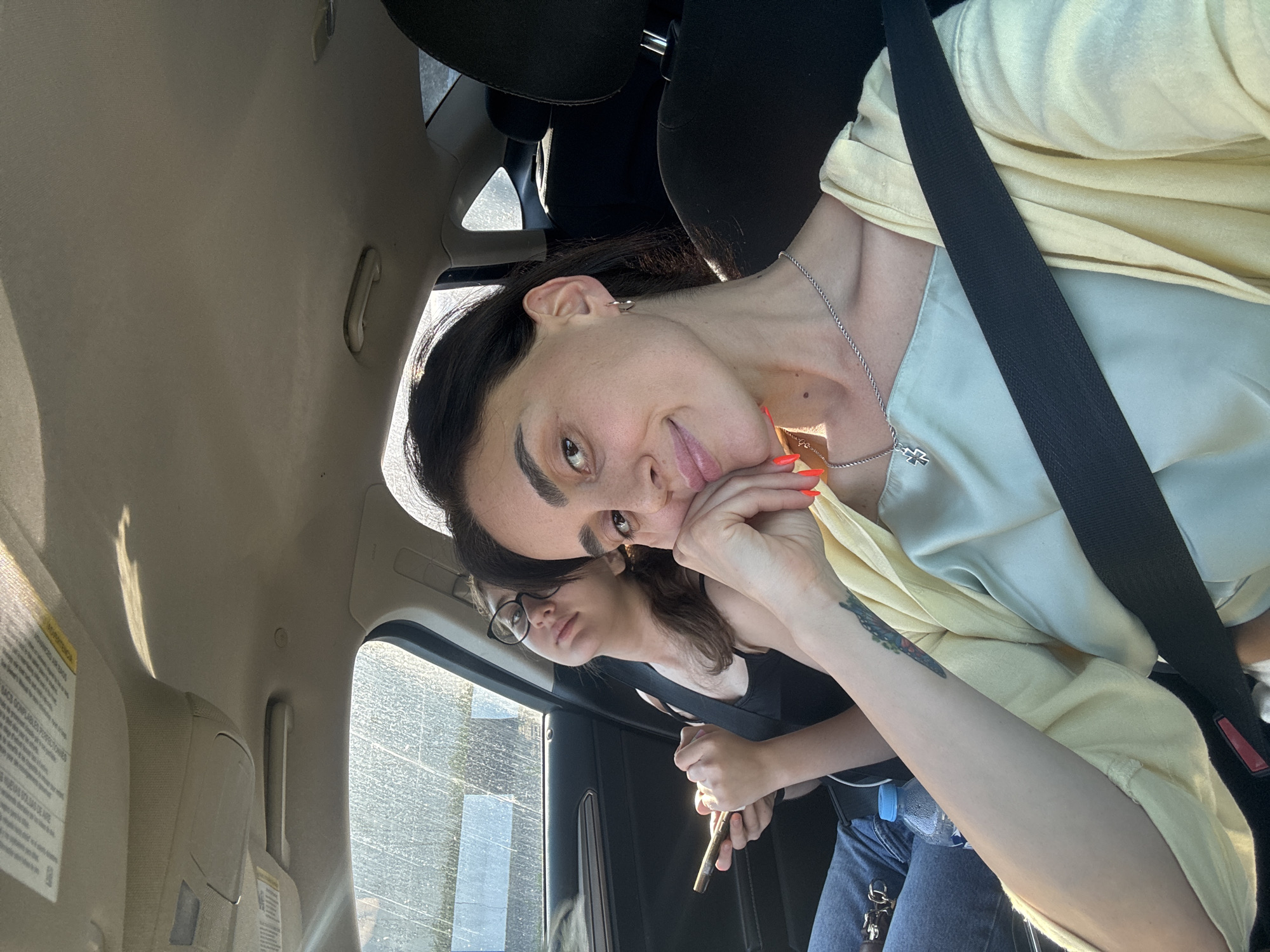 Two women are seated in the passenger seats of a car. The woman in the foreground, facing the camera, is resting her chin on her hand. She is wearing a light yellow open shirt over a light blue camisole and a silver necklace with a cross pendant. She has bright orange nail polish. A tattoo is visible on her left forearm. The woman in the background is wearing glasses and a black tank top. She is holding a smartphone in her left hand and looking up and to the left. A bottle of water is visible between the two women. The interior of the car is visible, including the dashboard, ceiling, and seats. The exterior of the car, seen through the windows, is blurred and appears to be a street scene with some greenery and sunlight. The image does not contain any specific visual information that would allow for the determination of the location in the city of Tbilisi, Georgia.