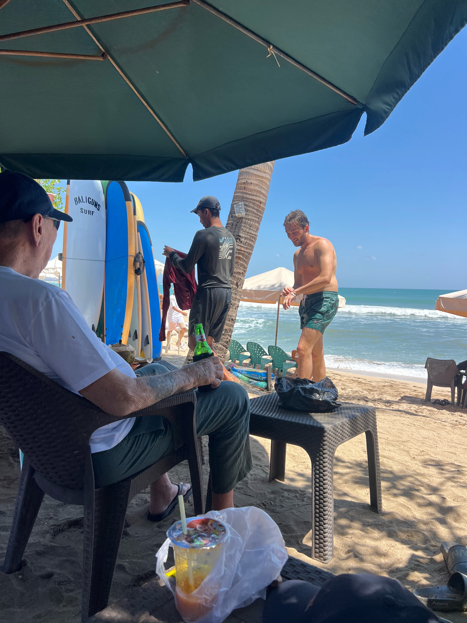 An overhead shot captures a beach scene in Kuta, Indonesia, under a large green umbrella. In the foreground, a man wearing a white t-shirt and dark pants sits in a dark plastic chair, holding a beer bottle with a koozie in his right hand. A plastic cup with a colorful drink and a straw rests on a table in front of him. Behind him, several surfboards are propped upright, one with "BALIGUNS SURF" written on it. To the right, a shirtless man in dark green swim shorts stands near the ocean, looking down. Another man in the background, wearing a dark long-sleeved shirt and shorts, is seen holding a maroon garment. The sandy beach extends to the turquoise ocean with gentle waves under a clear blue sky. Umbrellas and plastic chairs dot the shoreline, and a palm tree trunk is visible on the left side of the frame.