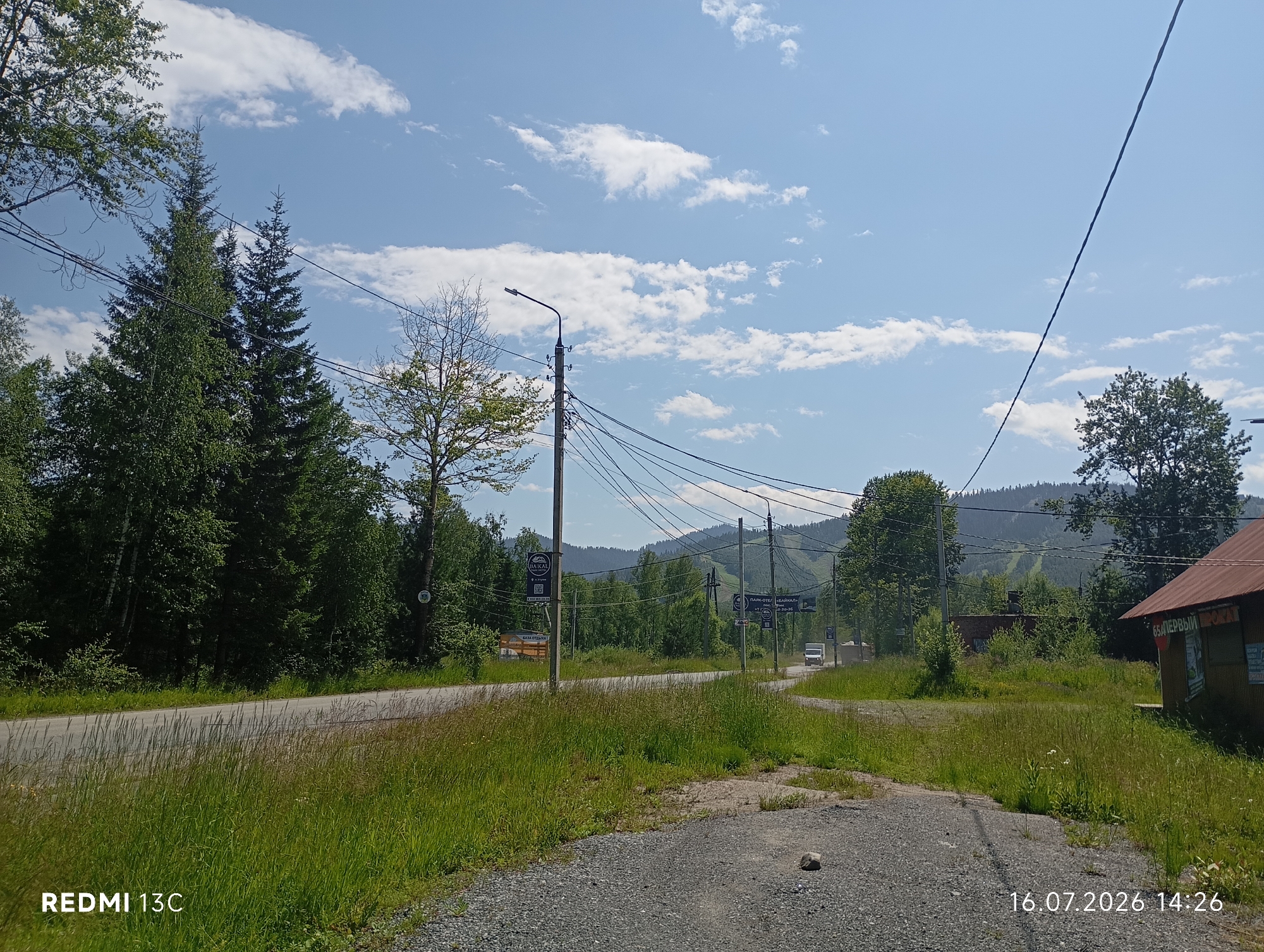 This image depicts a rural roadside scene in the vicinity of Baykalsk, Russia. The viewpoint is from a gravel path leading towards a paved road. On the left side of the road, a dense forest of evergreen trees dominates the landscape, with a few deciduous trees interspersed. A utility pole with numerous wires stretches across the frame, supporting a streetlight. On the right side of the road, a rustic building with a corrugated metal roof is visible, with some signage indicating it might be a shop. In the background, rolling hills covered in trees rise under a bright blue sky with scattered white clouds. A white van is seen driving on the road in the middle distance. The foreground is characterized by tall, green grass and gravel. The image is dated July 16, 2026, at 2:26 PM, as indicated by the timestamp in the lower right corner. The overall atmosphere is one of a quiet, natural environment on a sunny day.