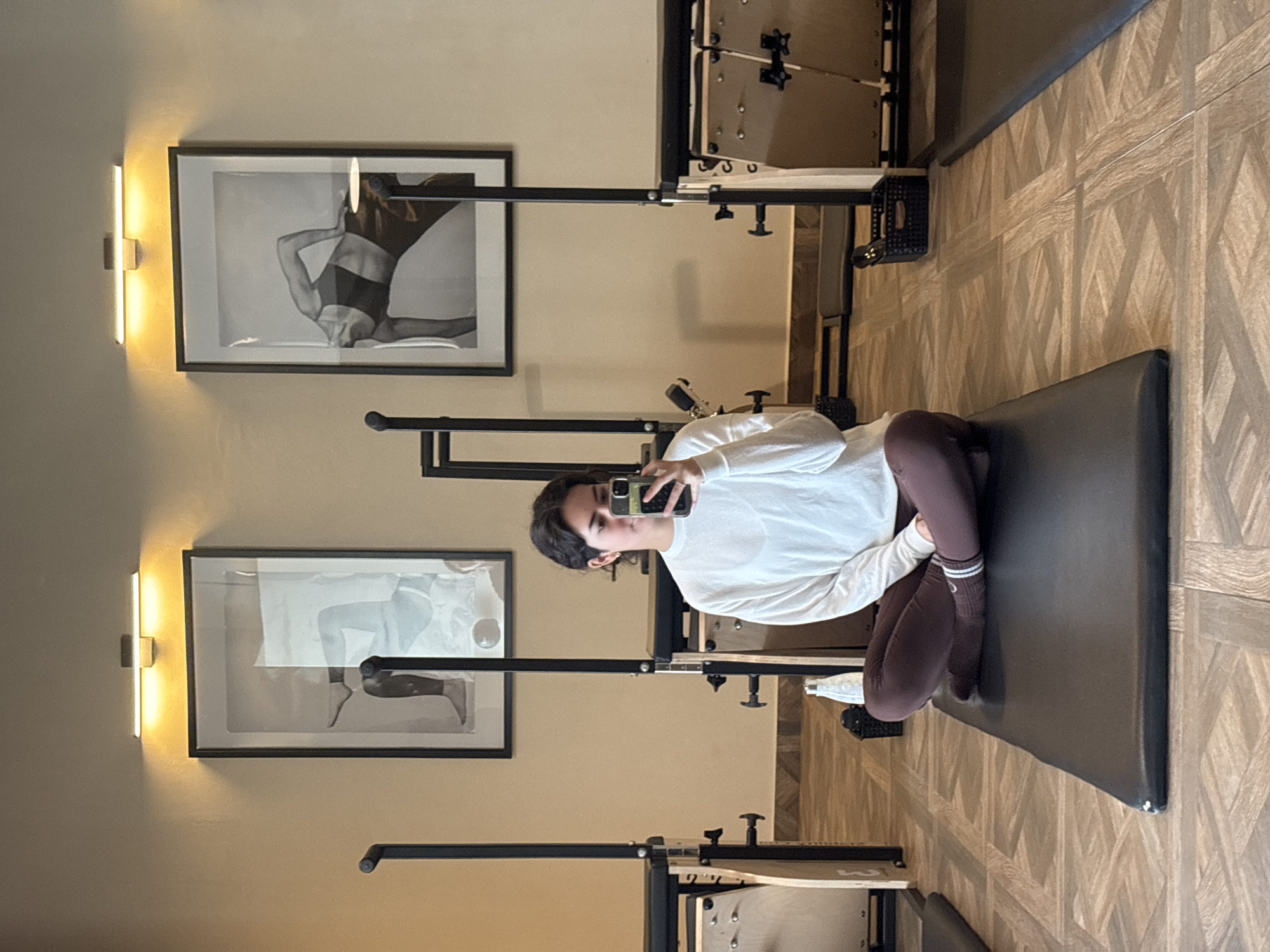 A young person in a white long-sleeved shirt and brown leggings is seated on a dark gray mat in what appears to be a pilates or fitness studio. The person is holding a smartphone and seems to be taking a selfie. Behind the person are two framed black and white photographs on a beige wall, each illuminated by a vertical light fixture. Pilates reformer machines are visible in the background, suggesting the room's purpose. The studio has a textured light brown and beige floor. The exact location in San Bernardino Tlaxcalancingo, Mexico, cannot be determined based on the given image.