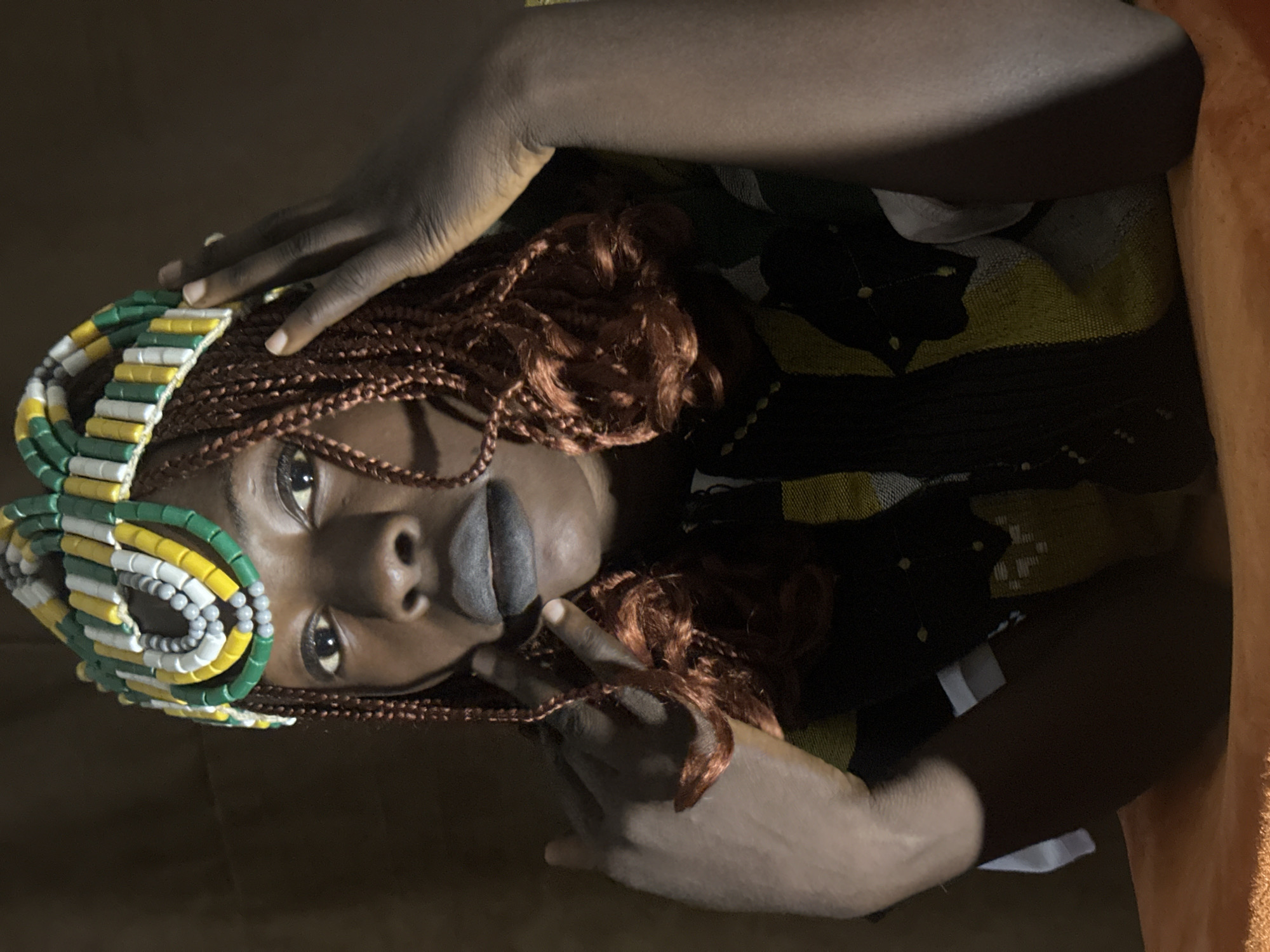 A young Black woman is shown from the chest up, wearing a beaded headdress and dark lipstick. Her hair is styled in reddish-brown braids that fall over her shoulders. She is wearing a brightly colored, patterned top. Her hands are raised, with one hand touching her hair near her temple and the other touching her chin. The background is a dark, textured surface, possibly a wall or fabric, and the foreground is a soft orange surface, likely a cushion or cloth. The lighting is dramatic, with highlights on her face and hands and shadows elsewhere. The scene suggests a portrait taken indoors. There is no visible indication of the city of Jos, Nigeria.