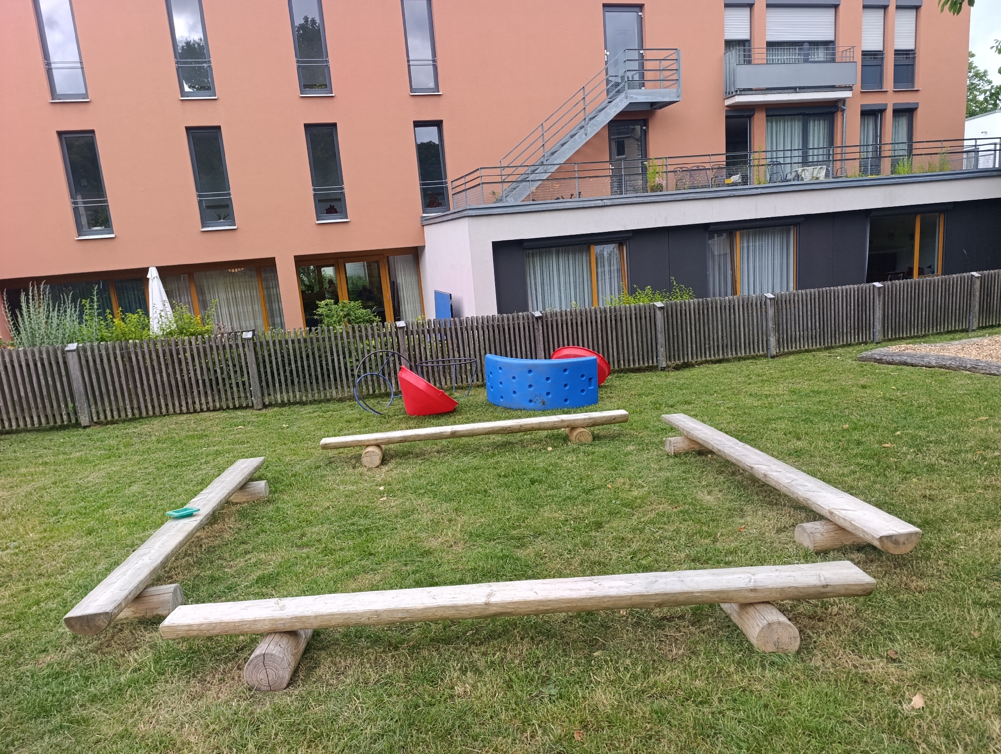 This image depicts an outdoor playground area located in the city of Pfullingen, Germany, in front of a modern, salmon-colored apartment building. The playground features several wooden balance beams of varying lengths and heights, constructed from rough-hewn logs. One longer beam is angled, resting on two smaller log supports, and has a small, turquoise object on its surface. Another, shorter beam is placed horizontally on two log supports, with a third beam positioned in front of it.
In the background, behind a weathered wooden fence, are some playground elements, including a red conical structure and a blue, circular object. The apartment building has numerous tall, narrow windows with dark frames, and balconies on upper floors. An external metal staircase leads up to one of the upper levels. The ground is covered with green grass, with some scattered fallen leaves. The overall scene suggests a relatively quiet, residential area.