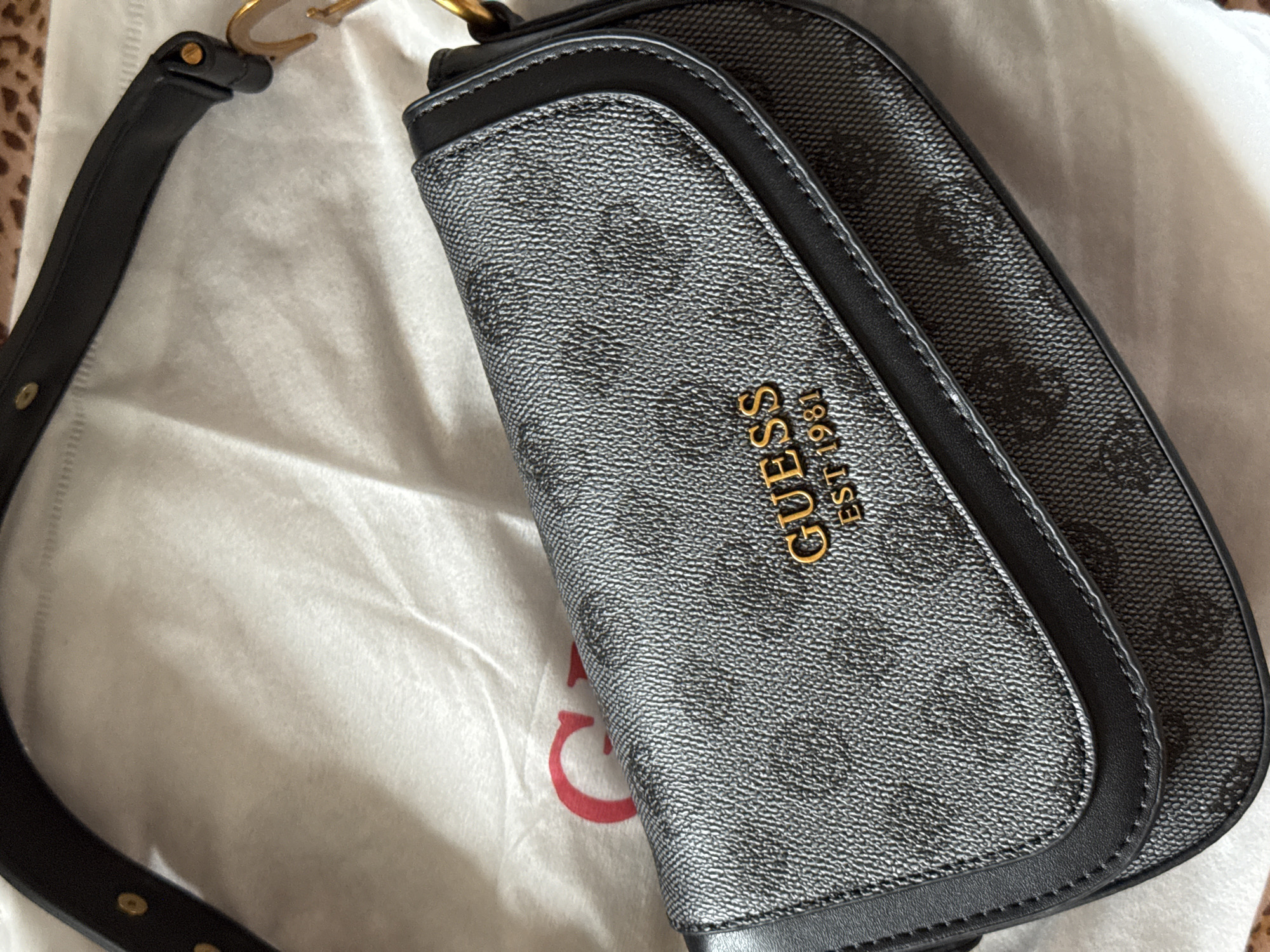 The image shows a close-up of a black Guess handbag with a subtle, repeating Guess logo pattern. The bag is made of textured material and has a black leather trim. Gold-colored lettering on the front of the bag reads "GUESS EST 1981". The handbag is resting on a white dust bag with a red "G" logo visible. The background shows a hint of a leopard print fabric. There is no indication of the location being Izhevsk, Russia.