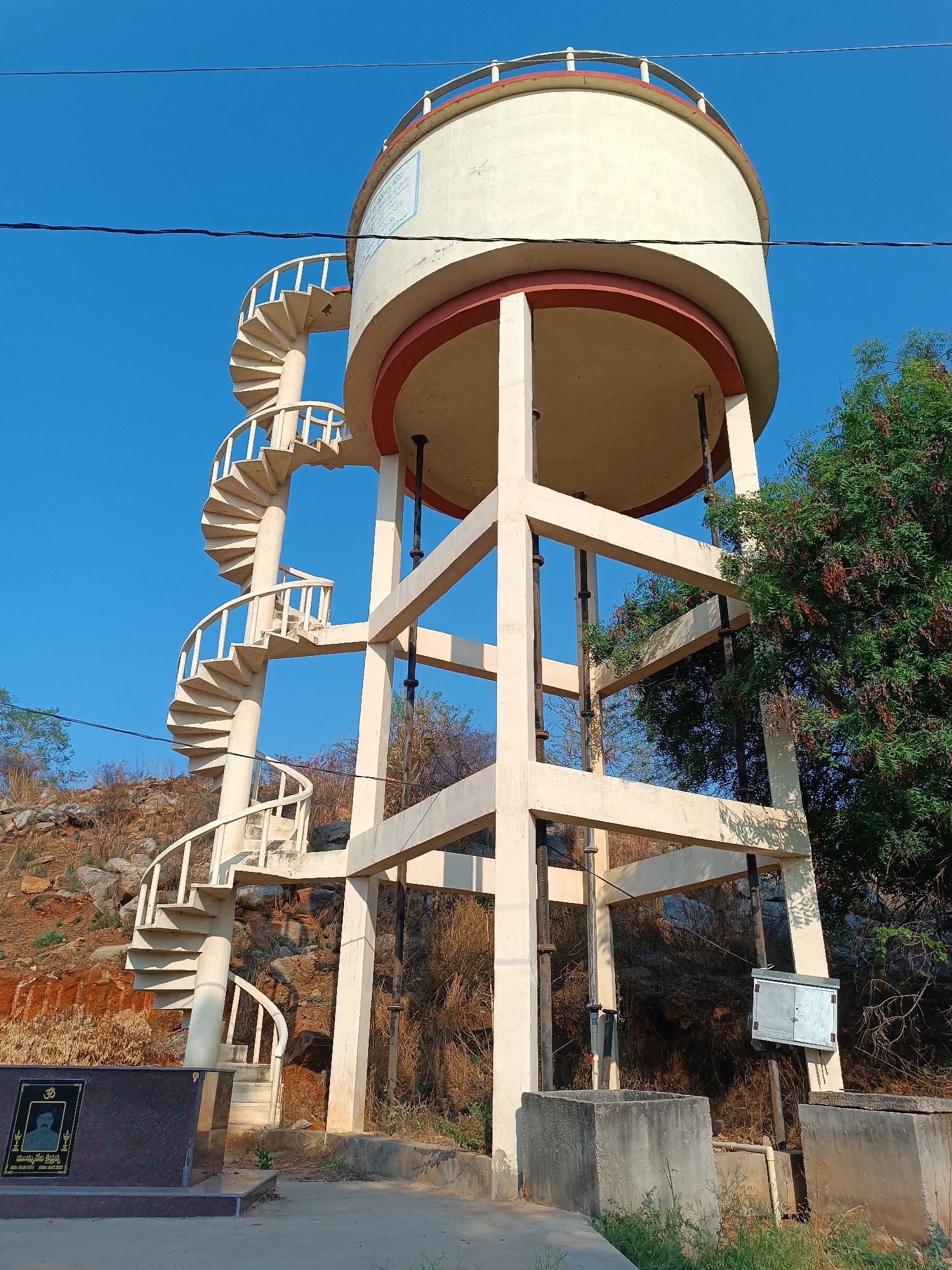 The image shows a water tower in Kammavaripalli, India, under a clear blue sky. The tower has a cylindrical water tank at the top, supported by a framework of square concrete beams. A spiral staircase wraps around one side of the tower, leading to the tank. Near the base of the staircase is a black granite memorial with a portrait on it. Behind the tower and staircase are rocky hills with sparse vegetation. To the right of the tower, trees partially obscure the view. At the base, there are concrete structures (possibly water intakes or outlets) and a metal utility box.