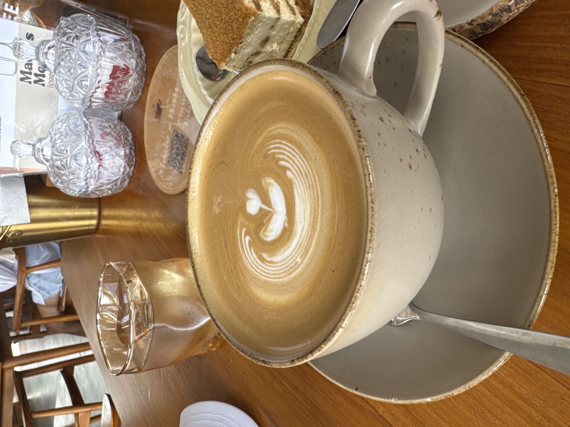 A close-up shot captures a latte with intricate foam art, served in a speckled ceramic mug on a matching saucer. The mug is positioned on a wooden table, with a glass of ice water to its left. In the background, two crystal jars with ornate lids contain what appear to be individually wrapped sweets. A slice of cake sits on a pale yellow plate to the right of the latte, accompanied by a spoon and a knife. The scene is set in a cafe, potentially in Kuta Selatan, Indonesia, suggested by the wooden furniture and the general ambiance of a tropical, relaxed setting.