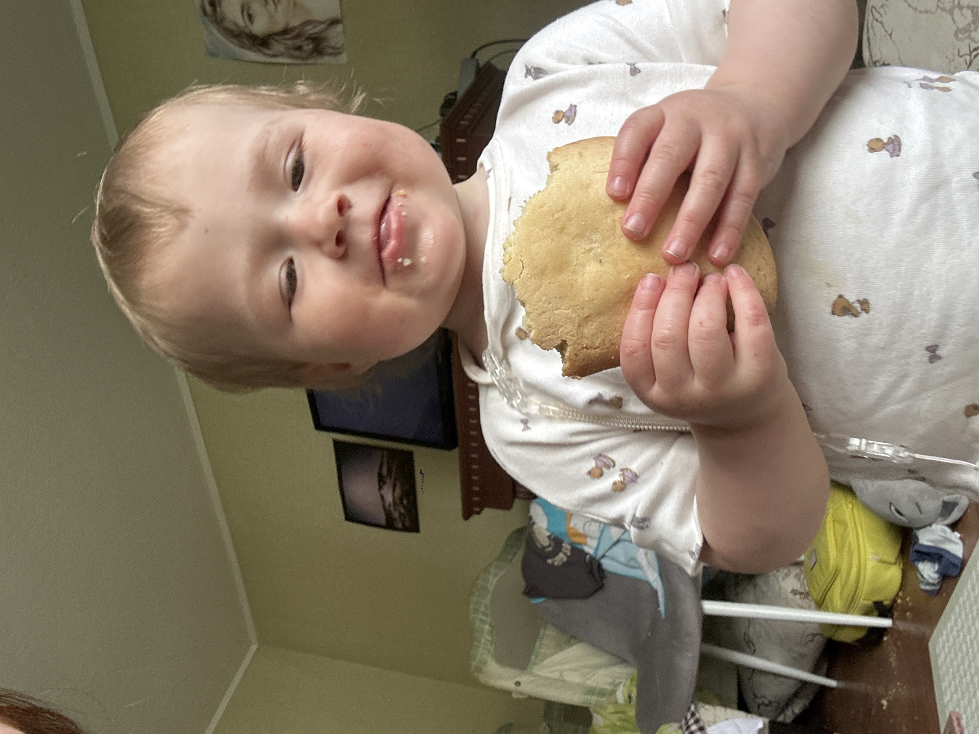 A close-up shot features a young child holding a partially eaten, round baked good (likely a cookie or pancake). The child has light skin, blonde hair, and appears to be around 1-3 years old. There is food residue visible around the mouth. The child is wearing a light-colored shirt or pajama top with small cartoon designs. In the background, there is a crib, a television, wall art, and hints of household clutter. The location cannot be determined from the information available. There is no data in the image that connects it with the city of Nizhny Novgorod, Russia.