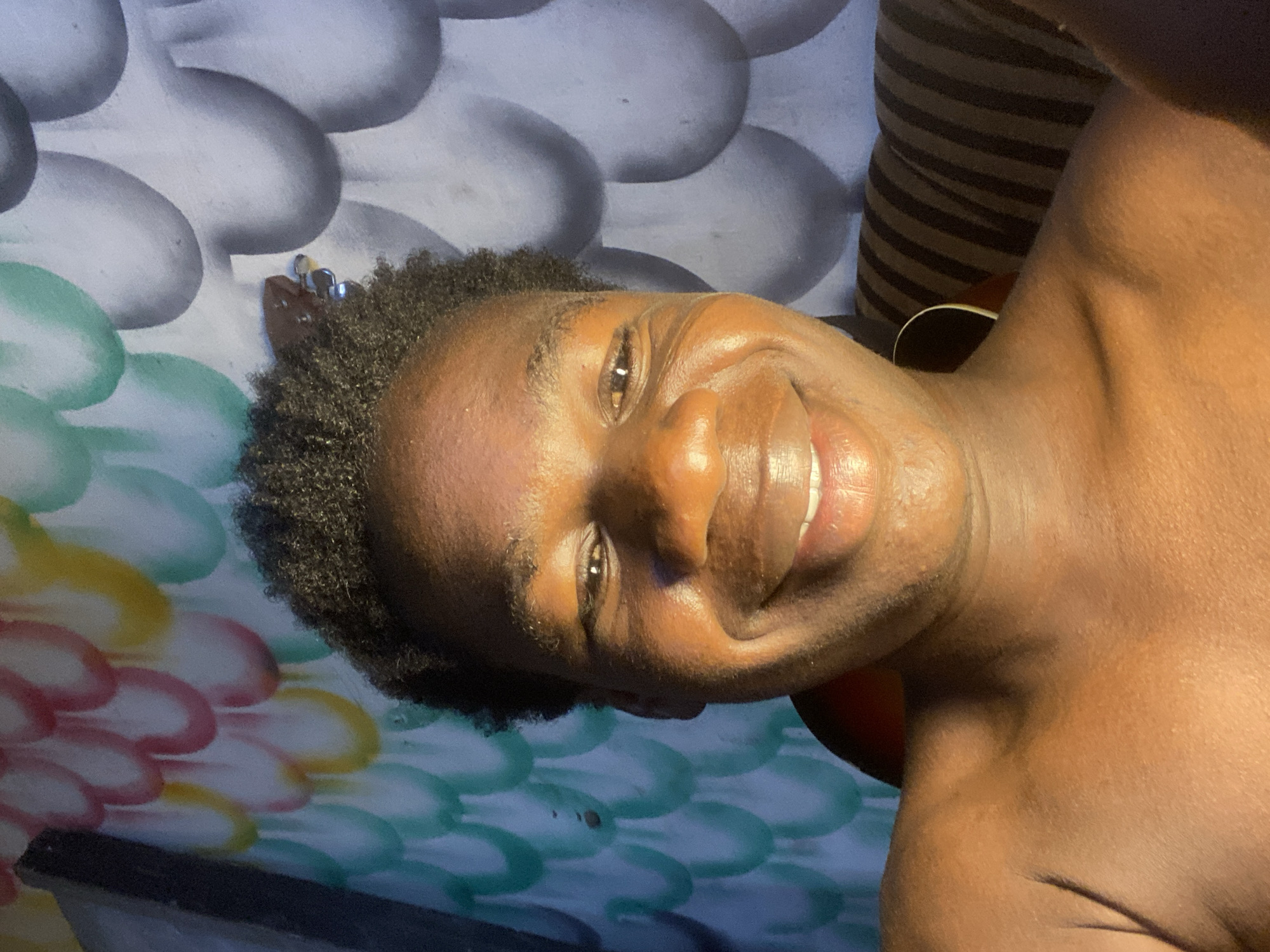 A close-up, rotated image shows a person with dark skin and short, dark, textured hair, smiling. The person's face is well-lit, revealing facial features like the nose, lips, and eyes. Part of the neck and upper chest are visible. The background includes a wall with a pattern of rounded shapes, some shaded in gray and others in colors such as green, yellow, and pink. There is also a striped object in the background. There is nothing in the image to indicate that it was taken in the city of Mararaba, Nigeria.