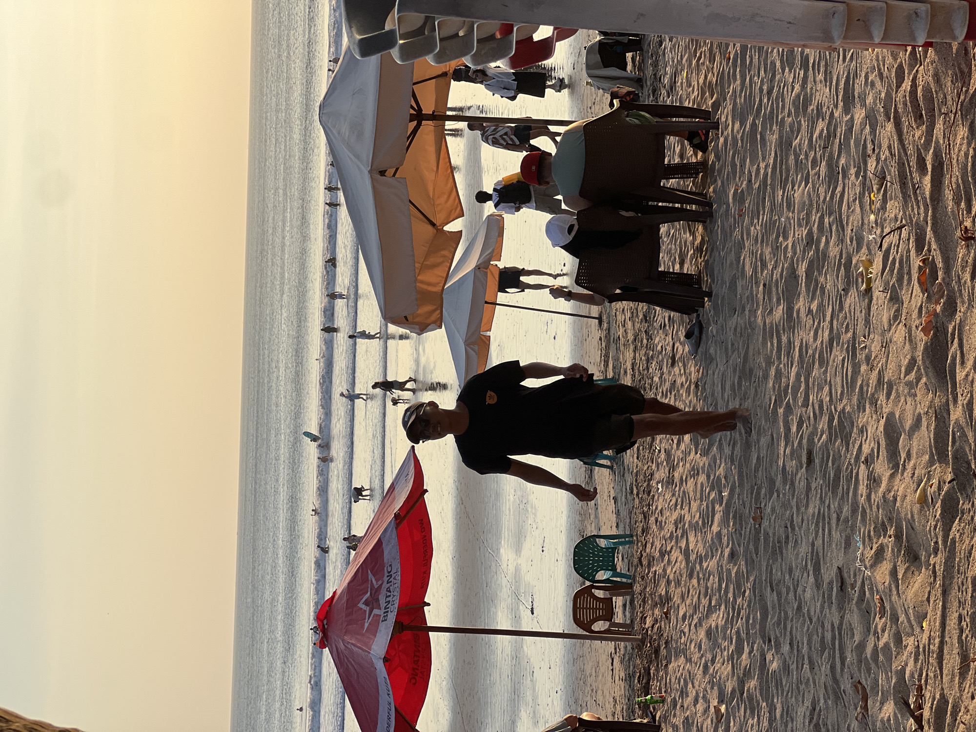 An outdoor, eye-level, medium shot captures a group of people on a beach in Kuta, Indonesia, at sunset. The sand in the foreground is golden and textured, with scattered leaves and debris. A man wearing a black t-shirt and shorts walks toward the viewer, his head turned to the side, with a baseball cap on. Behind him, two chairs sit empty under a large red umbrella. Further back, several people are visible in the shallow ocean, some with surfboards, enjoying the waves. Two larger, cream-colored umbrellas provide shade to groups of people sitting at tables. In the far distance, the ocean meets a hazy, pale orange sky.