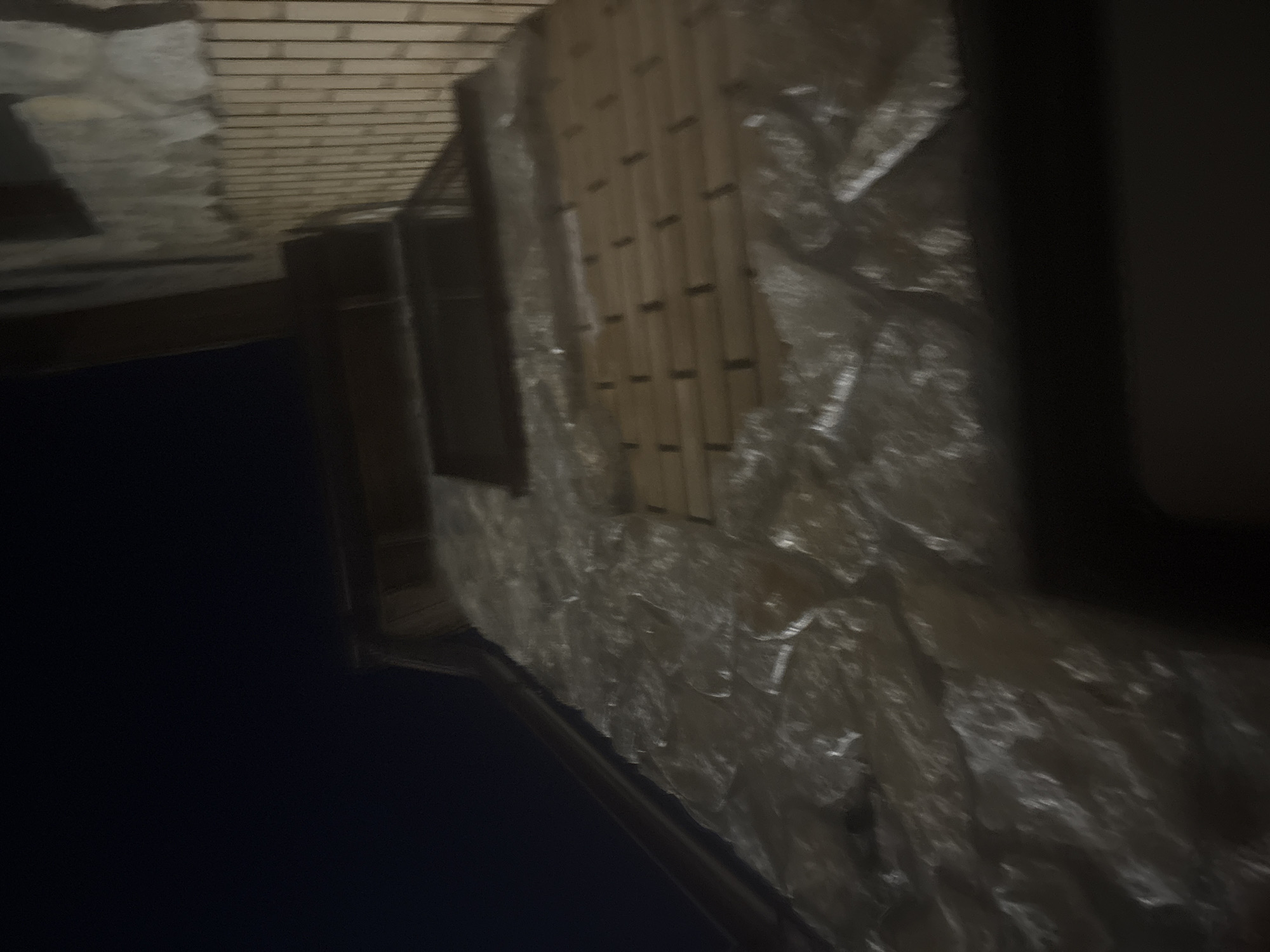 The image shows a close-up, low-angle shot of a building's exterior at night. The building appears to be constructed with stone on the lower portion and brick on the upper portion. There is a dark, possibly wooden, overhang or roof structure visible near the top of the frame. The stone facade is irregular and appears to be a rough-hewn, light-colored rock. The brickwork is composed of small, rectangular, light brown bricks laid in a horizontal pattern. A section of the brick wall appears to be recessed or a different material, with horizontal wooden slats or similar elements visible within the recess. The overall lighting suggests it is nighttime, with artificial light sources illuminating parts of the building, casting highlights and shadows. The background is a deep, dark blue, indicating a clear night sky. The specific location of Orekhovka, Russia, is not visually discernible from the provided image.
