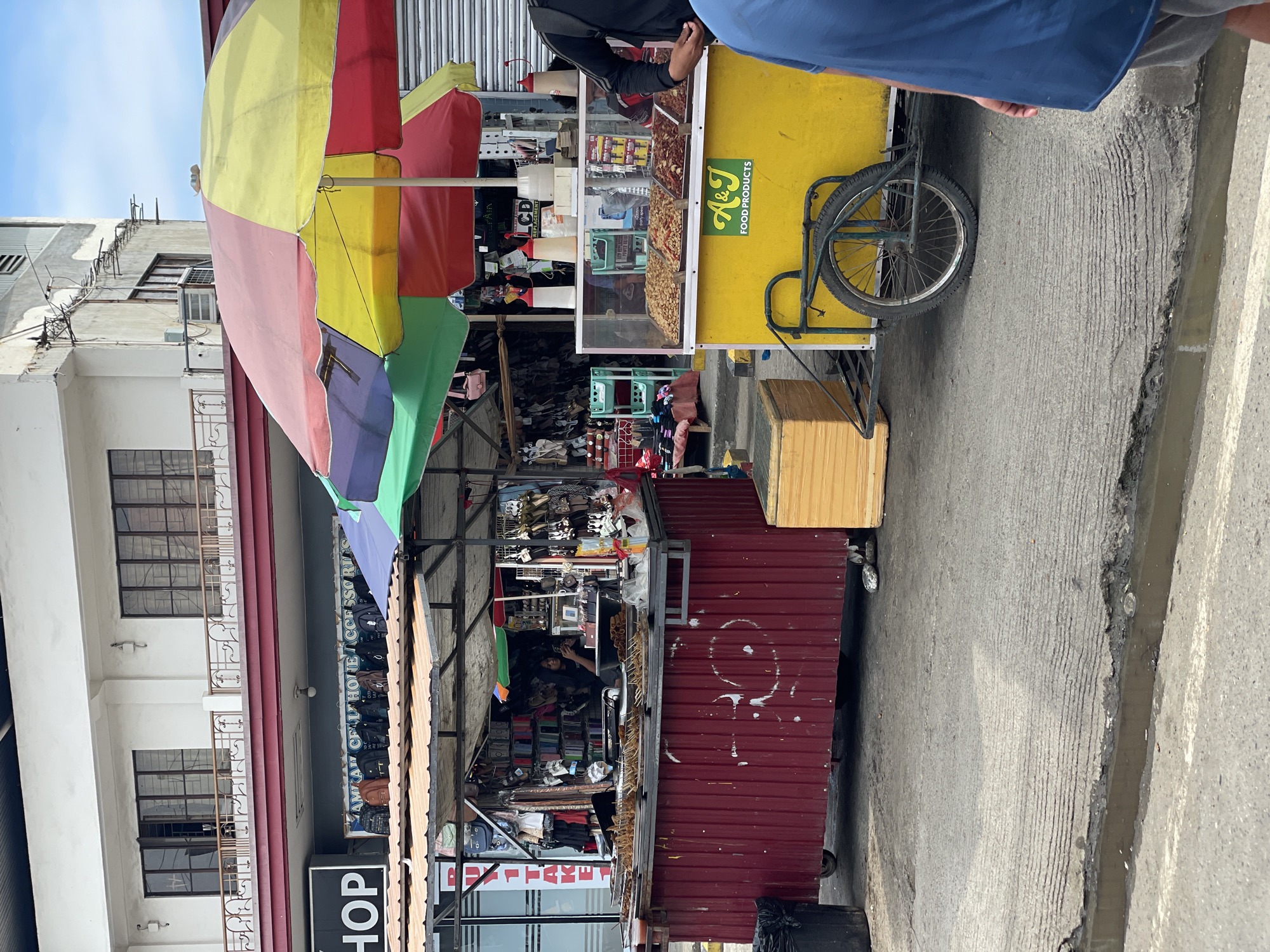 This image captures a daytime street scene in Davao City, Philippines, depicting a commercial area with vendors and a multi-story building.
In the background, a white two-story building features brown-framed windows on both levels and balconies with intricate dark metal railings. Above the lower-level shops, a sign partially reads "SHOP" on the left, and another sign further right displays "...AM AF CELL PHONE ACCESSORIES," with various bags hanging beneath it. Further to the right, another weathered, light-colored building with visible utility lines and an air conditioning unit is present. The sky above is clear and light blue.
In the midground, two primary street vendor setups are visible. On the left, a mobile stall with a corrugated red metal base and a dark metal frame offers items for sale. Trays of skewers, possibly grilled or fried food, are displayed on its counter. Inside the stall, a person with dark hair is seated, looking down at a mobile phone. A black trash receptacle is positioned to the left of this red stall.
To the right of the red stall, a bright yellow food cart, labeled "A&J FOOD PRODUCTS" in green text, is positioned. It features a transparent display case on top, showcasing three separate compartments filled with various dried snacks, appearing to be nuts or legumes. Two white squeeze bottles, presumably containing condiments, are placed on the cart's counter. A large, multi-colored umbrella (segmented in red, yellow, green, and blue/purple) shades this cart. The cart is attached to a bicycle frame, with one wheel visible in the lower right foreground. A light wooden crate or box rests on the ground next to the yellow cart on the left.
In the immediate foreground on the right, the lower torso and left arm of a person wearing a blue t-shirt and grey shorts are partially visible. The ground is concrete, textured with parallel grooves, and features a small, shallow puddle or channel of dark water running across the width of the frame at the bottom.
