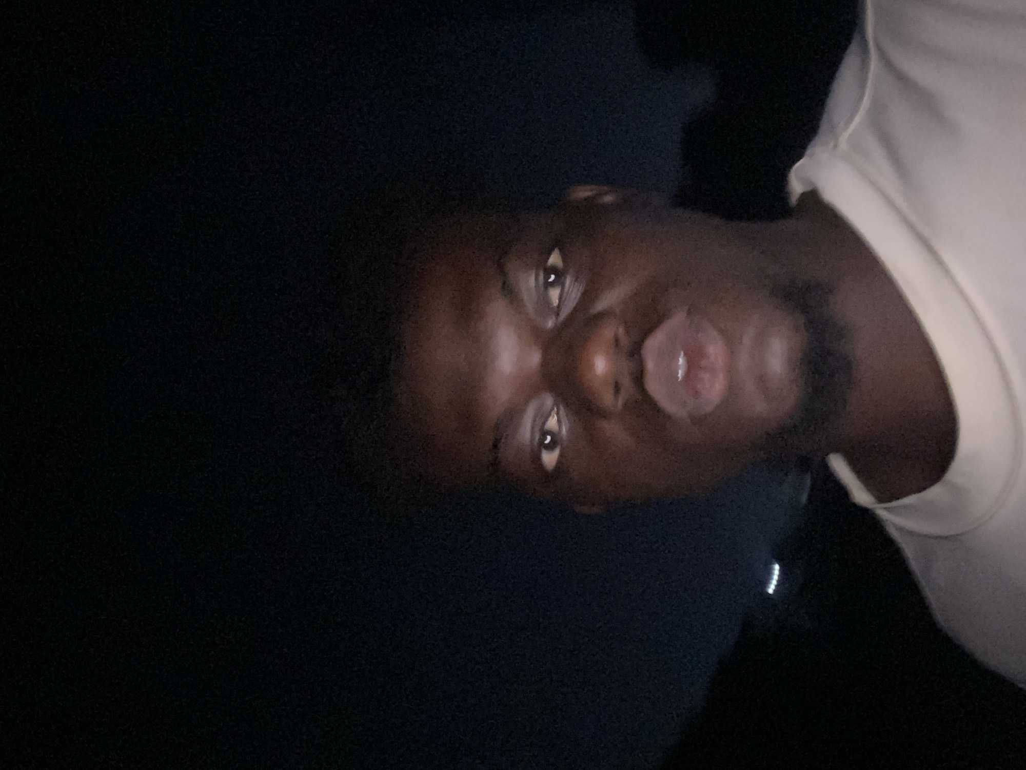 A man with dark skin and a beard is making a kissing face at the camera. He is wearing a light-colored shirt. The background is dark. The location is Unknown, Unknown.