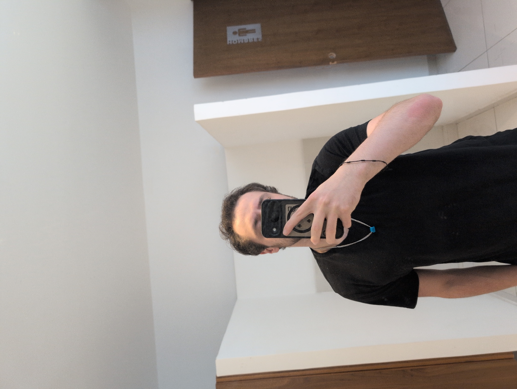 A medium shot captures a person taking a selfie in a restroom. The person is wearing a black t-shirt and a silver chain with a blue pendant. They are holding a black smartphone with a playing card design on the back, obscuring their face. In the background, a white partition wall is visible, as well as a brown wooden door with a "HOMBRES" (Men's) sign. The environment appears to be a public restroom. The image is cropped to show the person from the chest up, with their arm bent and hand holding the phone. A slight redness is visible on the person's elbow. The lighting is bright and diffused, suggesting overhead lighting. The overall scene is clean and functional.