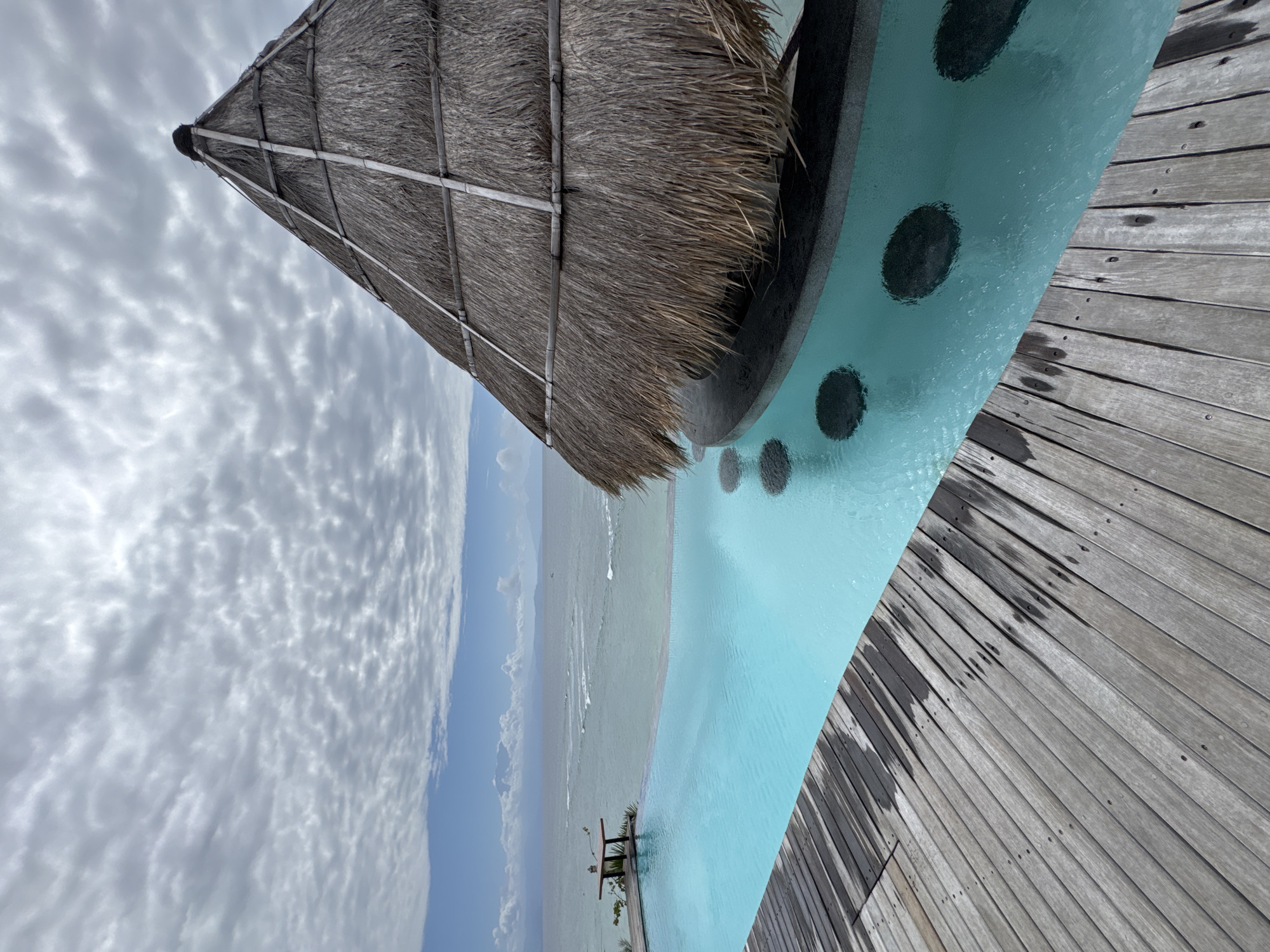 An outdoor infinity pool is located at a resort in Nusa Penida, Indonesia. The pool has bright blue water and is surrounded by a wooden deck. In the foreground, the wooden deck is wet in several places. A thatched-roof bar is situated in the pool, with several submerged circular seats visible. In the distance, the ocean stretches out to the horizon, with islands visible on the left. The sky is cloudy, with patches of blue sky showing through. There are no people visible in the image.