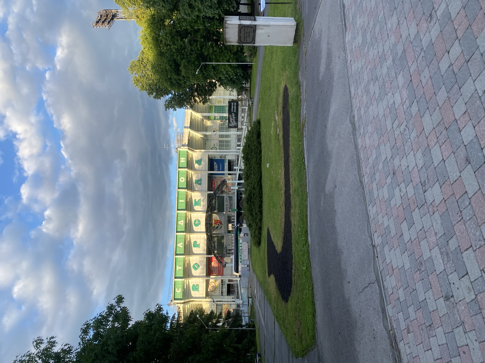The image shows a view of Vorskla Stadium in Poltava, Ukraine. The stadium's exterior is visible with vertical lettering "СТАДІОН ВОРСКЛА". There are advertisements for businesses like Ferrexpo, Fan&Sat, and Козацька Дроварня КРАФТОВЕ. The sky is partially cloudy, with sunlight illuminating the stadium. The image appears to have been taken from a pathway or sidewalk, next to a grassy area with a black-colored design. To the right is a paved area made up of red and gray bricks.