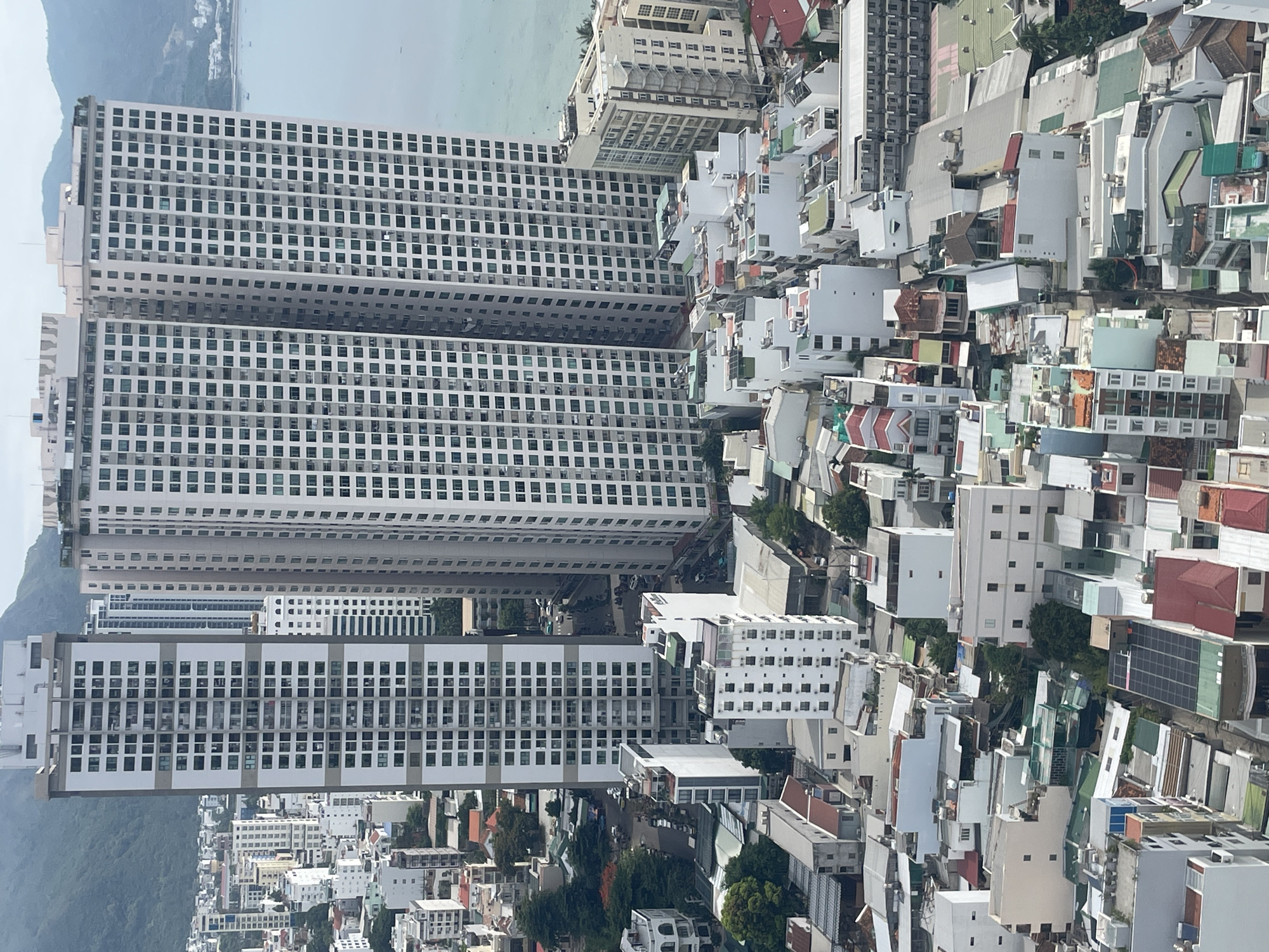 This is a high-angle, aerial shot of a dense urban environment in Nha Trang, Vietnam, likely overlooking a coastal area. The dominant feature is a cluster of tall, modern apartment buildings with repetitive window patterns. In the foreground and middle ground, the scene transitions to a more varied collection of smaller buildings, many with flat roofs and some appearing to be residential. Rooftop details include air conditioning units, water tanks, and some solar panels. Streets are visible weaving between the buildings, with a few parked vehicles. In the background, a body of water, likely the ocean, is visible alongside distant, hazy mountains. The overall impression is a bustling city with a mix of high-rise development and lower-density housing, situated near a natural coastal landscape. There are no discernible people or specific actions captured in this image.