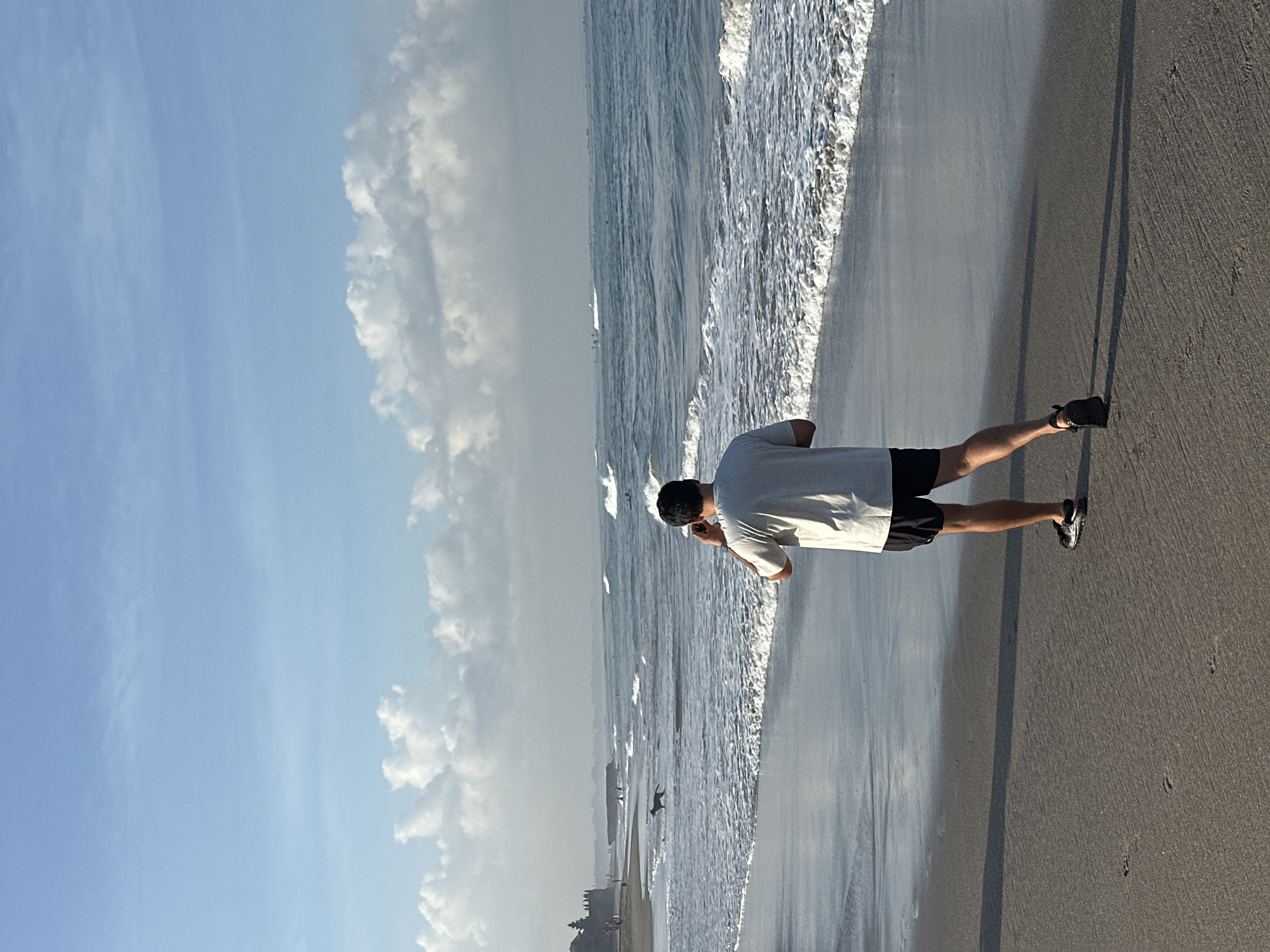 A person stands on a sandy beach at the edge of the ocean. The person is wearing a light-colored t-shirt and dark shorts. Their back is to the camera as they look out at the water. The ocean has small waves breaking on the shore, and there are some birds in the distance. The sky is blue with some fluffy white clouds. The sun appears to be low in the sky, casting long shadows on the sand, suggesting it is either early morning or late afternoon. The scene is peaceful and serene. The location is North Kuta, Indonesia. There is no visible text in the image.