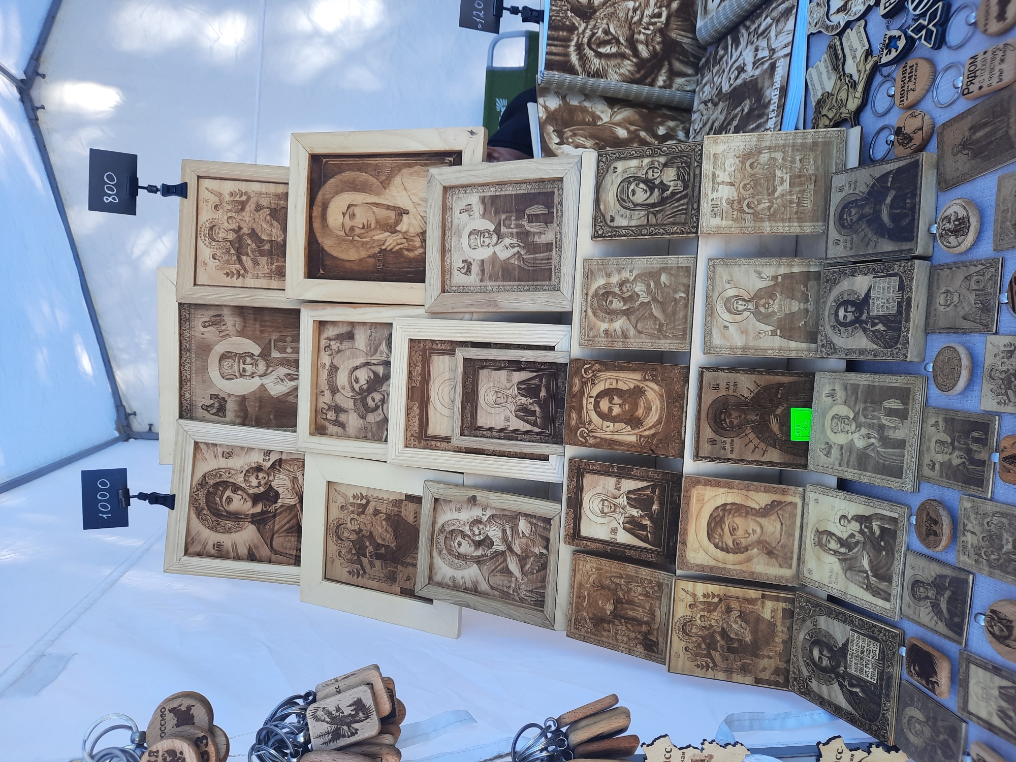 This is a street market scene in Zelengorsky, Russia, featuring a display of religious icons and wooden crafts. The primary subject is a tiered arrangement of framed wooden pyrographic art, predominantly depicting Christian imagery such as the Virgin Mary and Child, Jesus Christ, and various saints. The items are displayed on white fabric-covered surfaces, likely tables or stalls, under a white tent or awning. Several price tags are visible, with handwritten numbers "1000" and "800" on black signs. To the left, a collection of wooden keychains with pyrographic designs, including eagles and Cyrillic text, hangs from rings. In the background, a person wearing dark clothing and possibly a green garment is partially visible, suggesting a vendor or shopper. The overall environment appears to be an outdoor market on a sunny day, with the tent providing shade.
