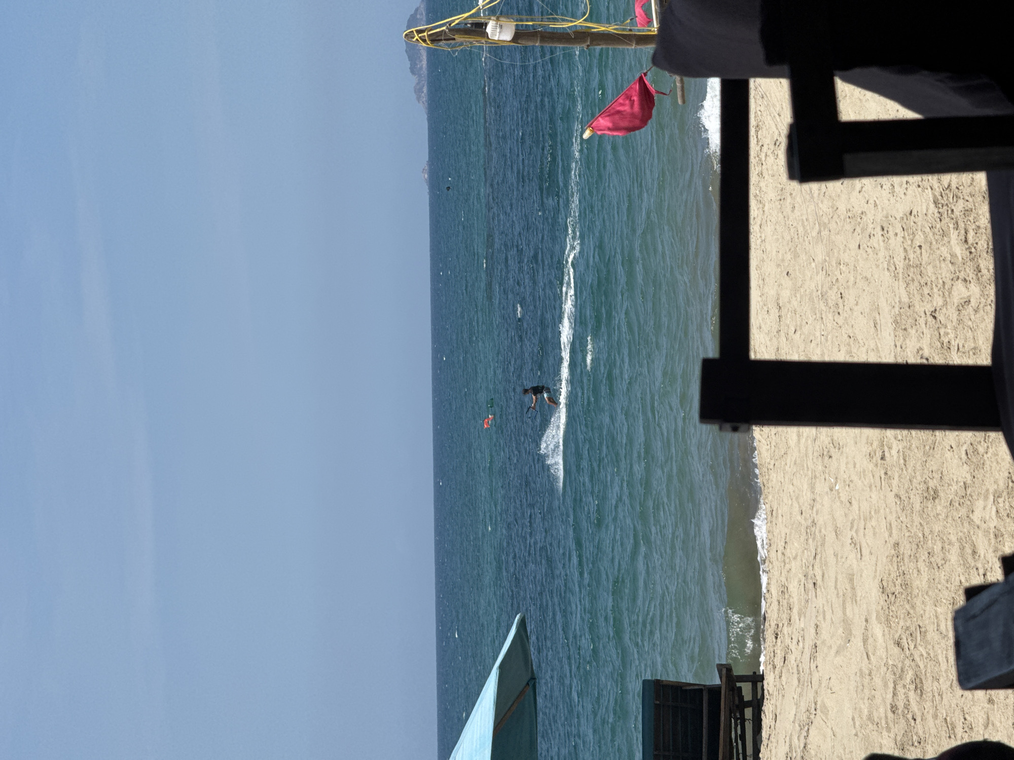 This is an outdoor scene taken at a beach in Đà Nẵng, Vietnam. In the center of the image, a person is kite surfing in the ocean. A red flag is visible behind them. The shoreline is to the right, with sand and surf meeting the water. An obscure object is situated in the lower portion of the foreground. The sky is clear and blue.