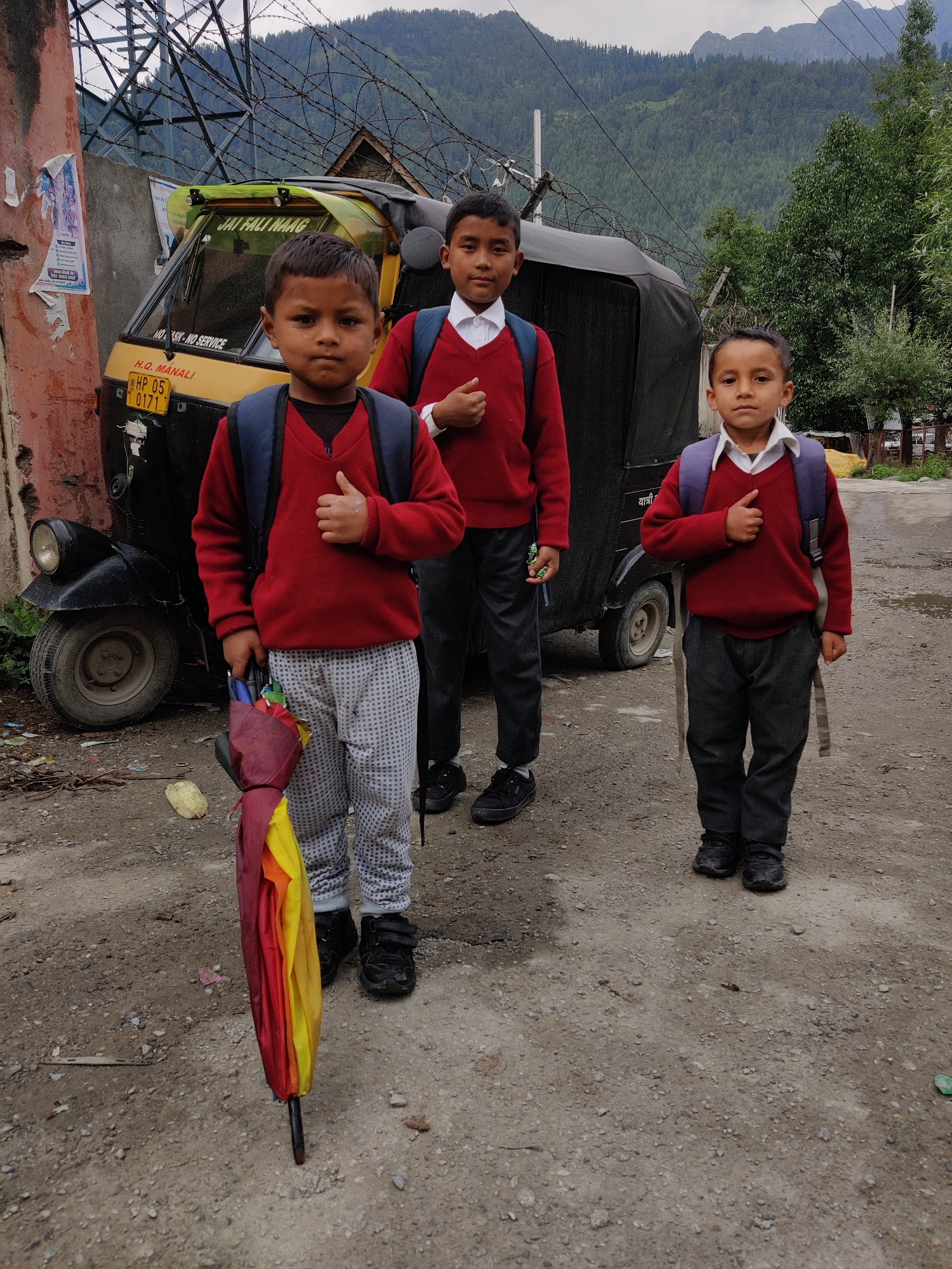 Three young boys, likely students, stand on a dirt road in the city of Bahang, India. The boy on the left holds a closed, multi-colored umbrella. All three boys wear red sweaters over white collared shirts and dark pants, with backpacks on their shoulders. In the background, a yellow and black auto-rickshaw is parked, and a barbed wire fence runs across the scene. Beyond the fence, a dense forest covers the hills, with mountains visible in the distance under an overcast sky. The ground is covered with dirt and small stones, with some debris scattered around.