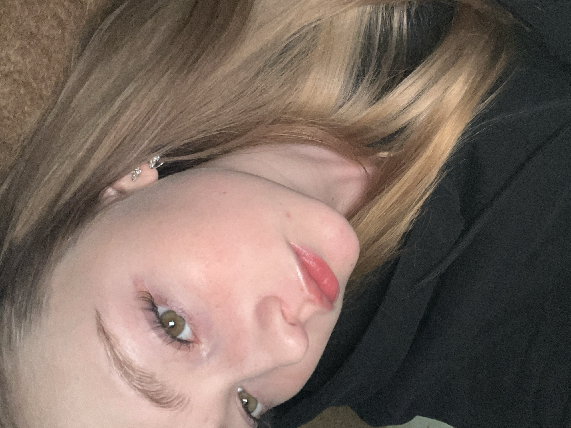 A close-up, angled portrait depicts a young woman with light-colored, highlighted hair. She has fair skin, light eyebrows, and hazel eyes. Her lips appear glossy, possibly from lip gloss. She is wearing an earring in her left ear. She's wearing a black garment, possibly a hoodie or sweatshirt, in the lower and right parts of the image. The background in the upper left shows a textured surface, possibly fabric or a sofa. The location is not discernible from the image; there is no indication the image was taken in Chaikovsky, Russia.