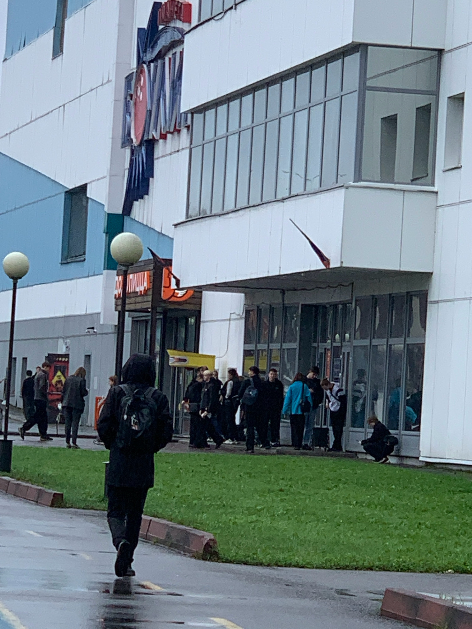A street-level, medium-shot, eye-level photograph captures the exterior of a multi-story building in Prokopyevsk, Russia, on a wet, overcast day. The building's facade is predominantly white with sections of light blue. Prominently displayed on the upper portion of the building is a large, red and blue sign with Cyrillic lettering, appearing to be a business or brand name. Large, modern windows dominate the upper sections of the building, reflecting the sky. Below these are a series of glass doors and windows at ground level, leading into the building.
In front of the building, a patch of bright green grass is bordered by a reddish-brown curb. Several streetlights with spherical tops stand on the left side of the image. A prominent sign with orange lettering and a cartoonish mascot is visible on the right side of the building entrance.
A diverse group of individuals are present in front of the building, some walking, others standing in small groups, and one person is crouched down. The foreground features a paved pathway that is wet from rain, with puddles reflecting the overcast sky.
A person wearing a black hooded jacket with a backpack walks away from the camera towards the building entrance. Other individuals, dressed in various outerwear, are seen near the entrance, some interacting with each other. The overall atmosphere is one of casual activity in an urban setting, with the wet conditions suggesting recent or ongoing rainfall.
