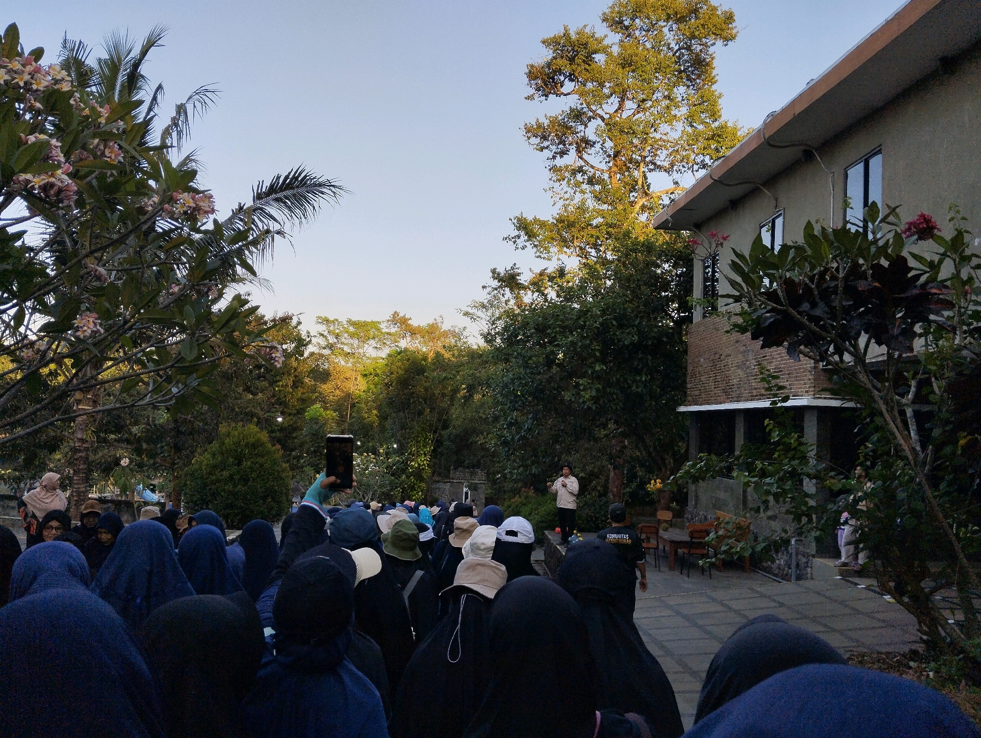 In Trisobo, Indonesia, an outdoor gathering of people is taking place in front of a building with a light-colored textured facade and a tiled roof. A large tree with lush green foliage stands tall to the right of the building, its branches extending towards the clear blue sky. In the foreground, a large group of individuals wearing dark blue and black hijabs are assembled, many looking towards the center of the scene. Some people are holding up mobile phones, possibly to take pictures or record videos. To the left, flowering trees with pink and white blossoms add a touch of color to the environment. Further back, a man in a light-colored shirt and dark pants stands on a raised platform, appearing to address the assembled crowd. He is gesturing with his hands, suggesting he is speaking. Behind him, more trees and foliage create a natural backdrop. The ground is paved with gray tiles, indicating an outdoor public space. The overall atmosphere appears to be a communal event or educational session taking place in a tranquil, green setting.