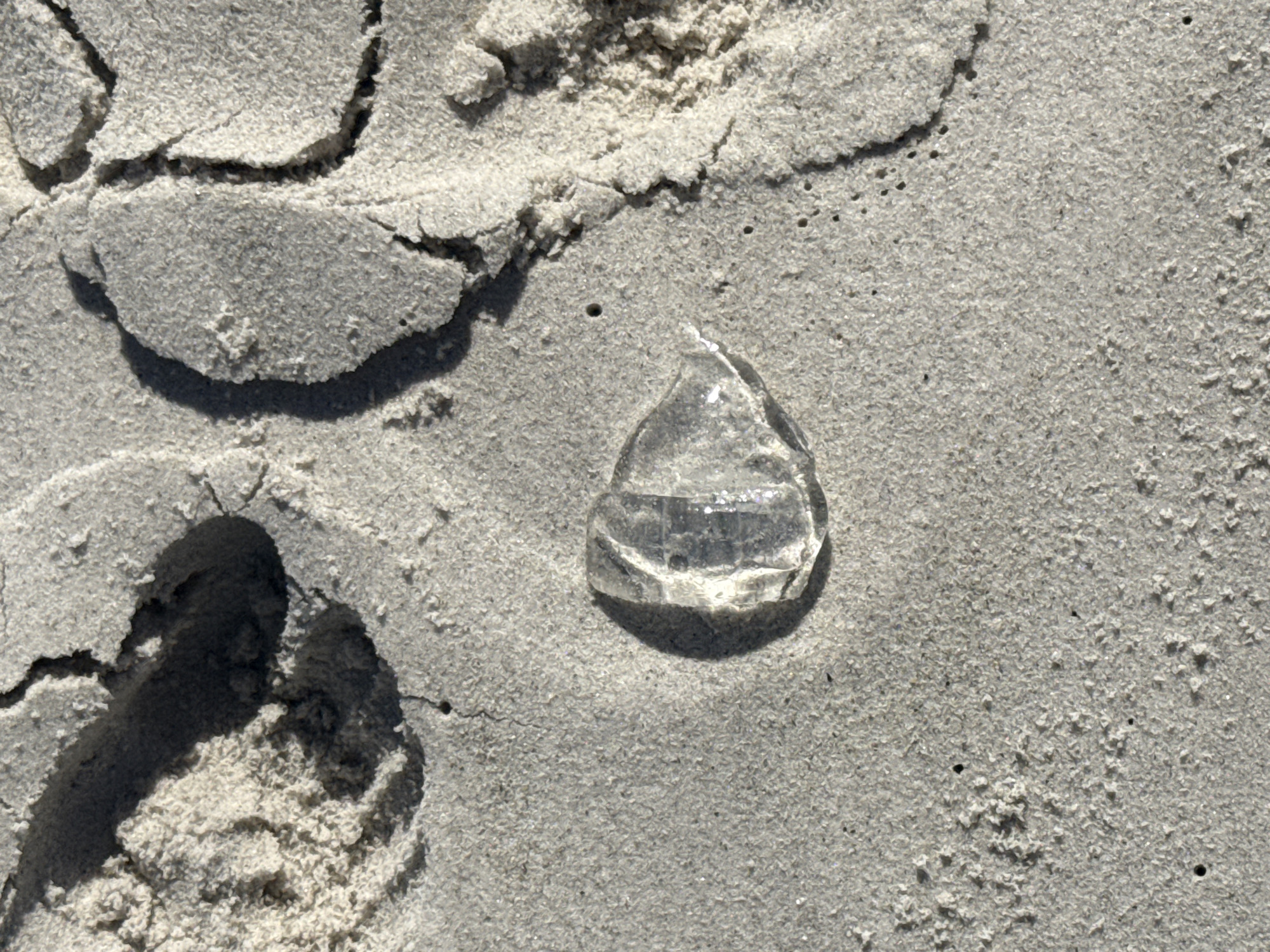 The image depicts a section of sandy beach, likely in Cape Town, South Africa. The sand is light grey and fine-grained. A piece of clear glass, roughly teardrop-shaped, sits on the sand surface, partially buried. Footprints are visible in the sand, displaying an indentation and disturbed area, indicating recent human activity. Small, dark dots are scattered across the sand surface, possibly indicating small organisms or dark mineral grains. The overall scene is one of tranquility and discarded refuse on a beach environment.