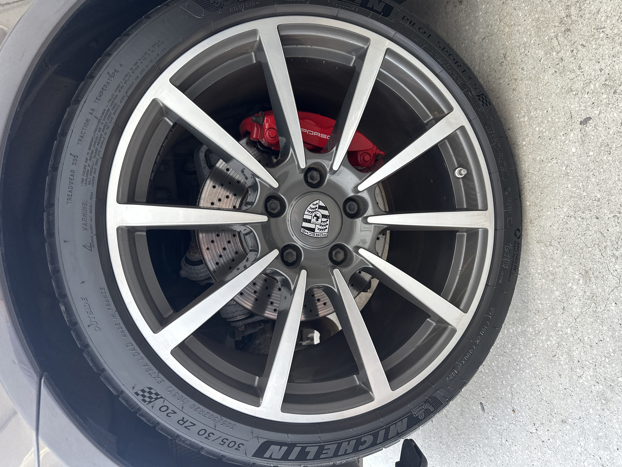 The image shows a close-up of a Porsche wheel and tire, likely taken in an outdoor setting given the concrete ground. The tire is a Michelin Pilot Sport 4, with specifications "305/30 ZR 20" visible. The wheel is a multi-spoke alloy design with a gunmetal gray finish and polished accents. Prominently featured are the red Porsche brake calipers. The tire's sidewall displays various markings, including "OUTSIDE," "MADE IN FRANCE," "TREADWEAR 300," "TRACTION AA," and "TEMPERATURE A." There are also warning labels regarding tire safety. The car's bodywork is visible at the top, a dark gray color. The scene is static and focuses solely on the wheel and tire assembly.