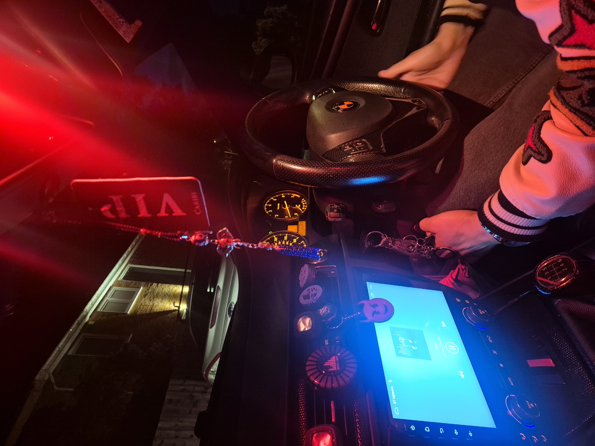 A person sits in the driver's seat of a car at night. The car's dashboard is visible, including the steering wheel, gauges, a large touchscreen display emitting a bright blue light, and various buttons and controls. A "VIP" tag hangs from the rearview mirror. The person's hands are on the steering wheel and near the gear shift. A building is partially visible through the windshield. The scene is illuminated by interior car lights, including red and blue hues. The location in the city of West Suffolk, United Kingdom is indeterminable from the image.