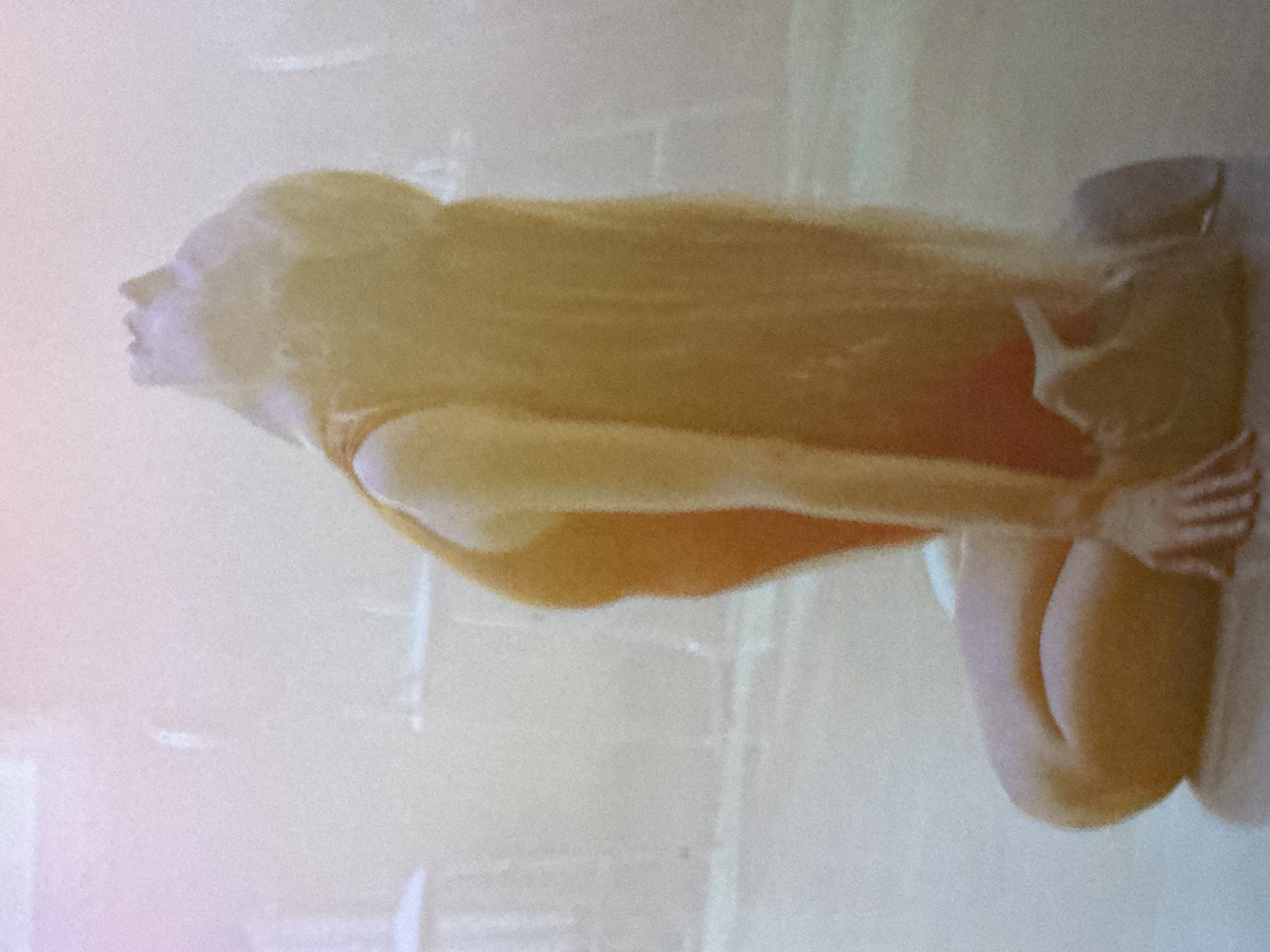 A person is kneeling on the floor with their head tilted back. They are wearing an orange leotard and their long blonde hair cascades down their back. The background is blurred and out of focus, with some indistinct shapes that could be furniture. The overall lighting is soft and diffused.