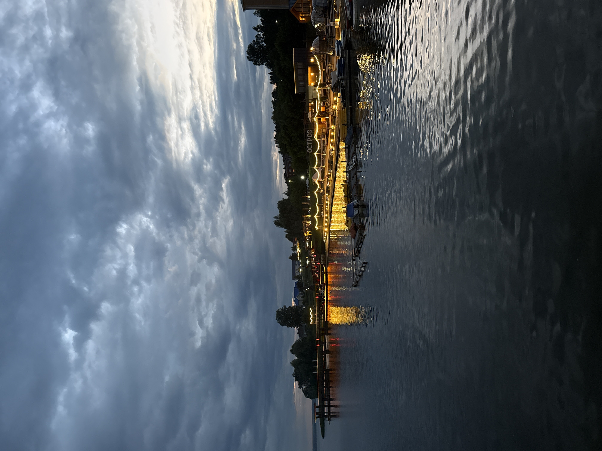 The image depicts a waterfront scene at dusk in Votkinsk, Russia. A calm body of water occupies the foreground, with gentle ripples reflecting the warm glow of lights from the shore. Along the shoreline, a wooden pier extends into the water, adorned with strings of festive lights. Several small boats are moored along the pier and further down the shore. In the background, a line of trees separates the waterfront from a cluster of buildings, some of which are also illuminated. The sky above is a dramatic canvas of dark, heavy clouds, tinged with the soft light of the setting sun peeking through. A sign with Cyrillic lettering, "ОСТРОВ" (Ostrov, meaning "Island"), is visible on the shore. The overall atmosphere is serene and inviting, suggesting a peaceful evening by the water.