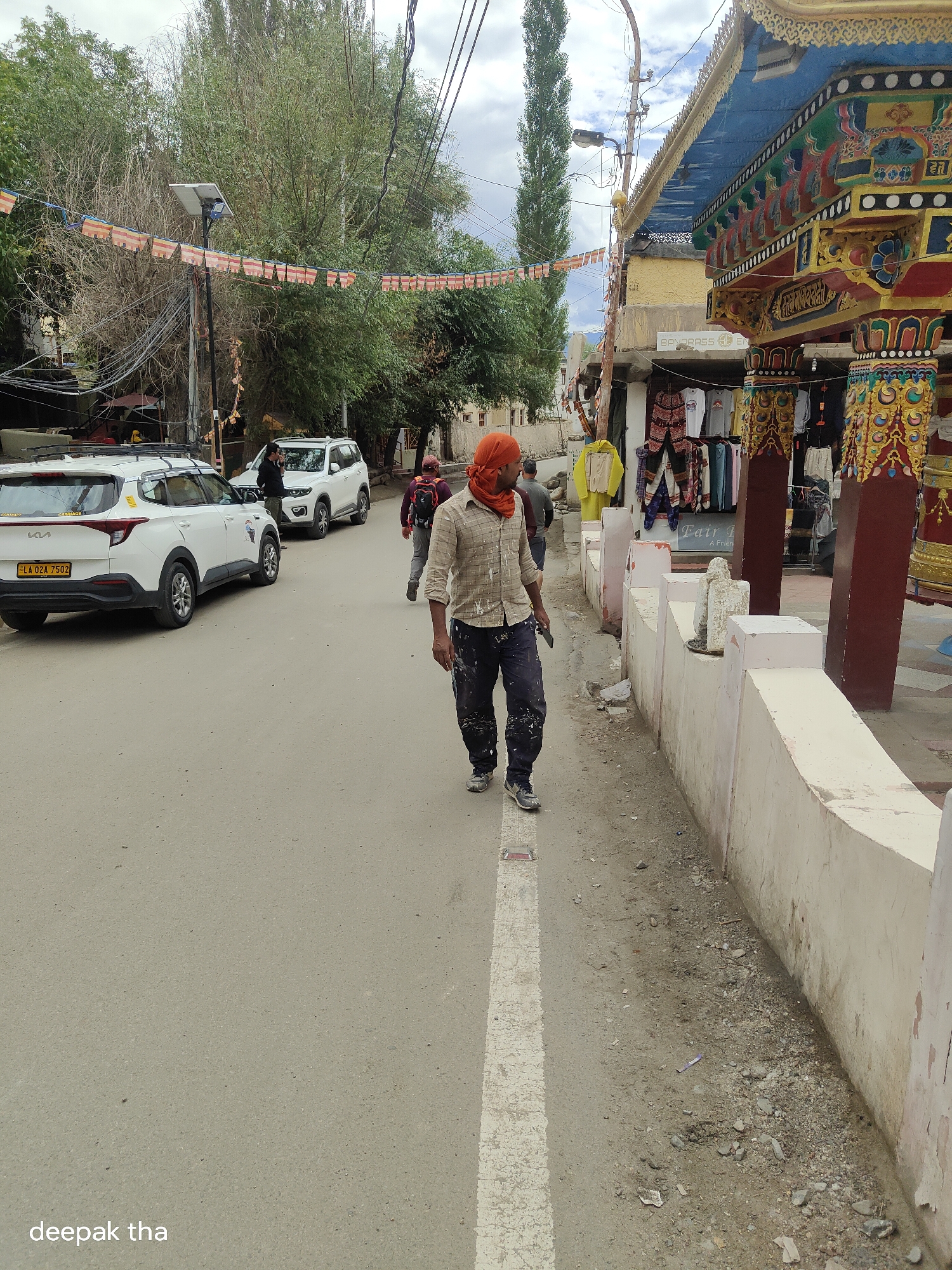 The image captures a street scene in Leh, India, likely near a Buddhist monastery given the decorative architecture and prayer flags visible. In the foreground, a man wearing an orange headscarf and a paint-splattered shirt walks on the asphalt road, holding a tool in his right hand. He is dressed in dark pants and athletic shoes. Behind him, two white SUVs are parked on the side of the road, and further back, another person is seen walking. The street is lined with buildings, one of which features colorful, ornate architectural details characteristic of Tibetan Buddhist style. Prayer flags are strung across the street, fluttering in the breeze. The environment appears to be a town or city street with buildings, vehicles, and pedestrians. The road has a white dividing line.