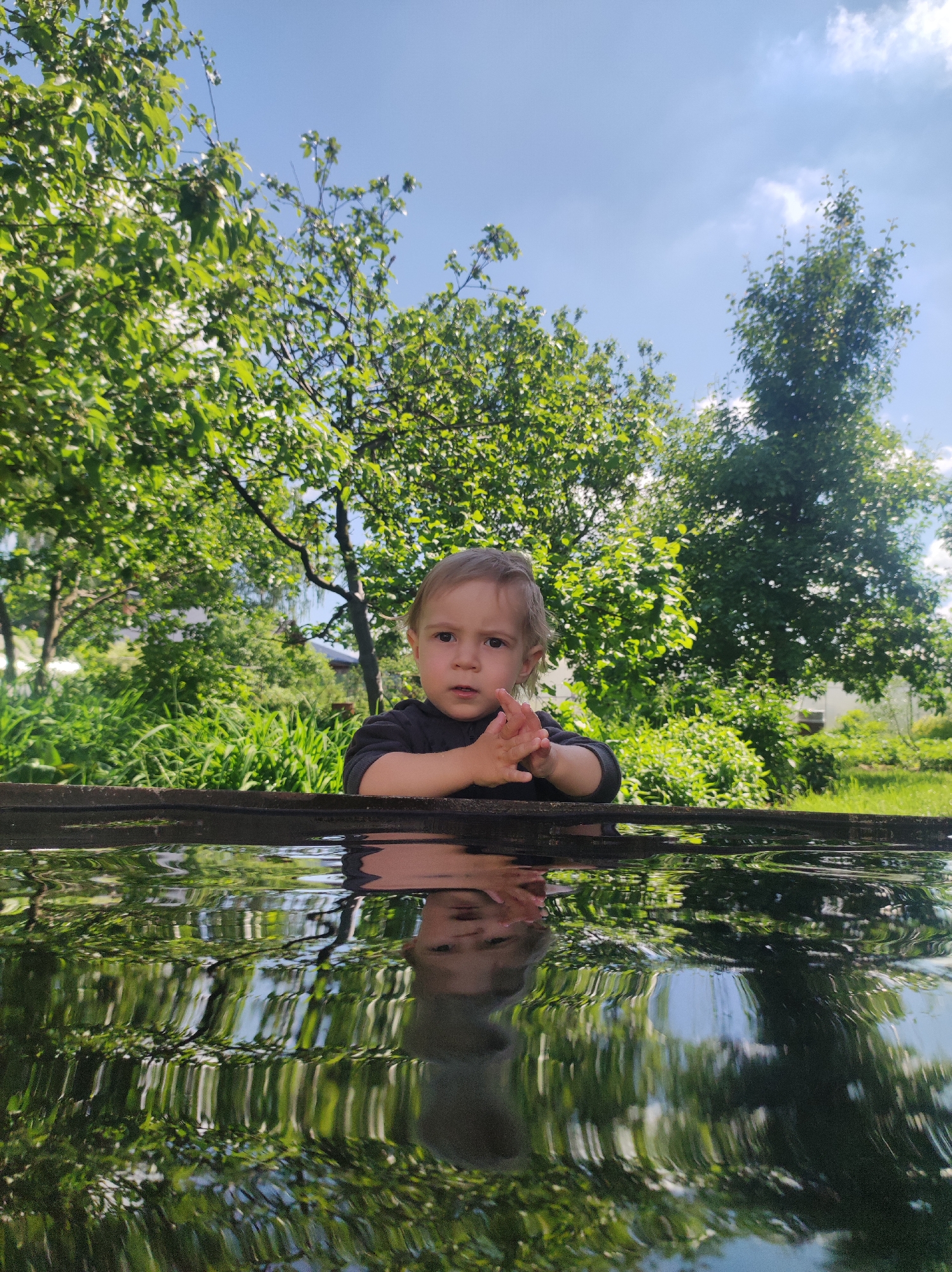 A young child, likely a toddler, is the central subject of this outdoor portrait. The child, with light brown hair and dark eyes, wears a dark long-sleeved top and looks directly at the camera with a neutral, slightly curious expression. Their hands are held together in front, appearing wet.
The child is positioned behind a dark, wooden-like trough or container filled with water, which occupies the lower half of the frame. The water's surface is rippled, creating a wavy and distorted reflection of the lush green trees and foliage in the background, as well as the bright blue sky above. Parts of the child's face and clothing are also visible in the reflection.
Behind the child and the trough, a vibrant outdoor garden or yard unfolds, dominated by numerous green trees and dense bushes under a clear blue sky dotted with scattered white clouds. The scene is brightly lit by natural sunlight, indicating a sunny day. The overall setting suggests a pleasant day in a natural, possibly rural or suburban, environment in Chaikovsky, Russia. There is no visible text in the image.