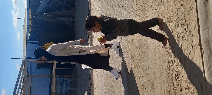 An outdoor scene in Khan Yunis, Palestinian Territory, features two individuals walking on sandy, unpaved ground under a bright blue sky with sparse clouds. In the foreground, a barefoot young child, dressed in dark pants and a dark grey hooded jacket, walks from left to right, holding a small, light-yellow object with both hands at chest level, looking towards the right. Behind and to the left of the child, an adult female walks in the same direction. She wears a black hijab, a long black dress, a light-colored woven cardigan, and white athletic shoes, with a white bag slung over her shoulder. Her profile is visible, facing forward. Long shadows of both individuals are cast on the ground to their left. The background shows improvised structures constructed from wooden frames, blue and dark tarpaulins, and possibly concrete blocks. A light blue plastic chair is visible to the left of these structures, and a partially obscured figure in a yellow top is present deeper in the background.