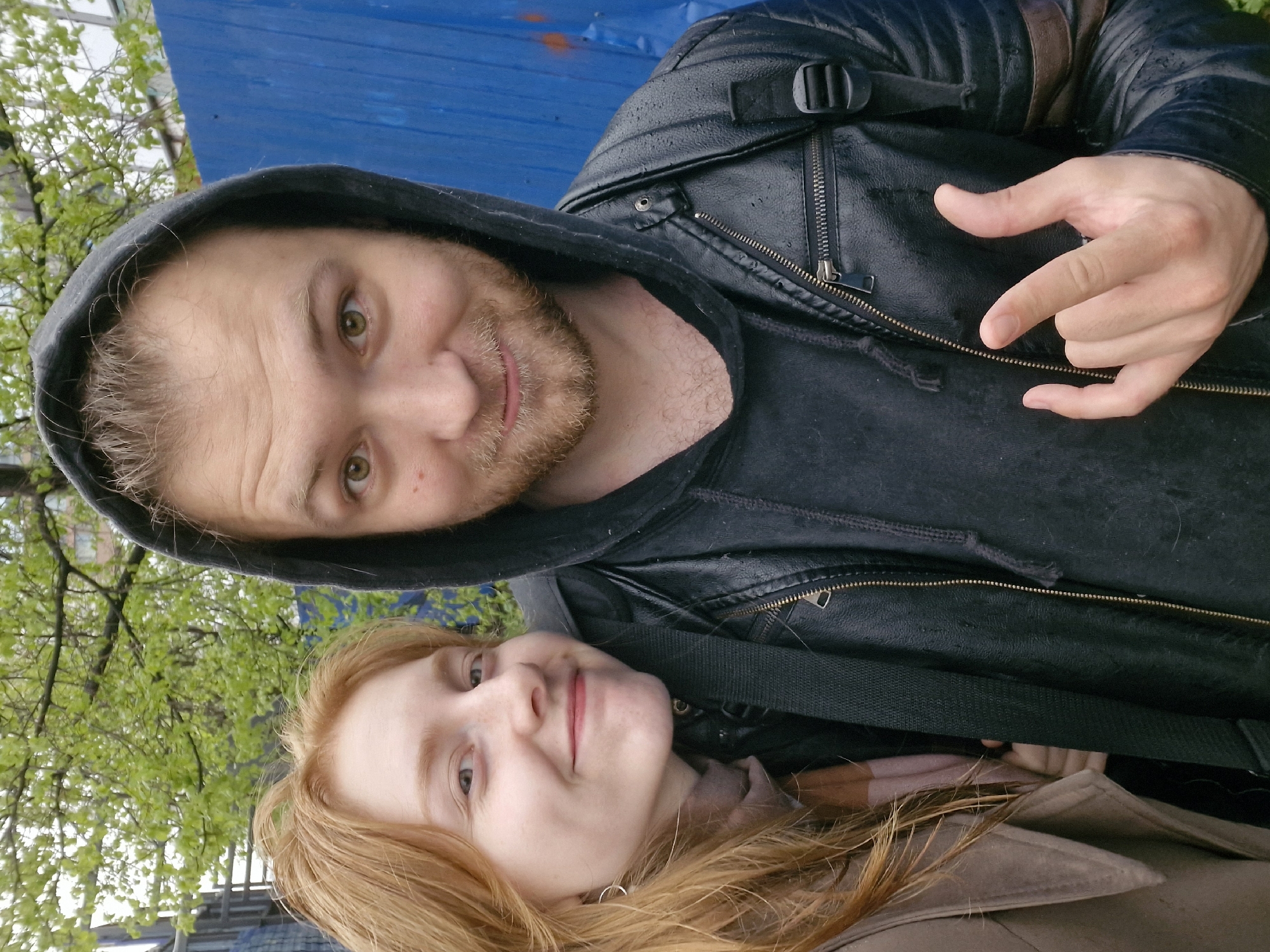 Two people are posing for a photo. The person on top is a man with light brown hair, a beard, and green eyes. He is wearing a black hoodie and a black leather jacket. His right hand is visible, making a hand gesture. The person below is a woman with long red hair. She is wearing a light brown coat. The background includes green foliage, a blue wall, and a bridge-like structure. Based on the architecture and greenery, this photo might have been taken outdoors in a city environment like Yekaterinburg, Russia. The image is slightly tilted.