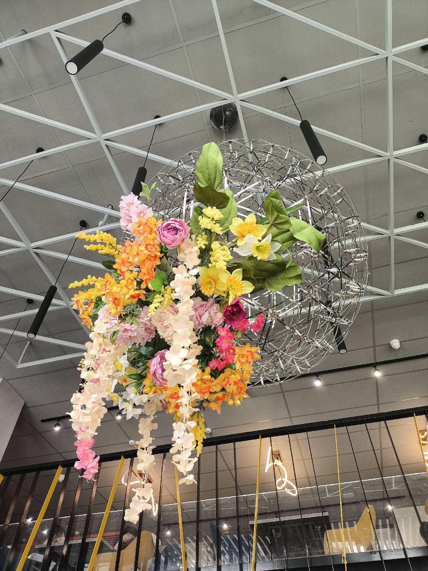 The image depicts an interior space, likely a commercial establishment in Izhevsk, Russia, given the presence of a hanging floral arrangement and modern design elements. The scene is viewed from a low angle, looking upwards.
The dominant feature is a large, spherical metal wire structure suspended from the ceiling. This structure is adorned with an abundant, colorful floral arrangement, featuring a variety of flowers in shades of pink, yellow, orange, and white. Green foliage is also incorporated into the arrangement. The overall effect is decorative and eye-catching.
The ceiling is composed of a gray, grid-like structure with white supports and black cylindrical lights. Below the floral arrangement and metal sphere, there's a black metal railing with yellow accents, suggesting a mezzanine or upper level. Behind the railing, there's a partial view of an interior space with chairs and a neon sign.
No people are clearly visible in the frame, but the presence of chairs behind the railing hints at occupancy. The overall ambiance is modern and stylish, likely within a restaurant, cafe, or retail space.
