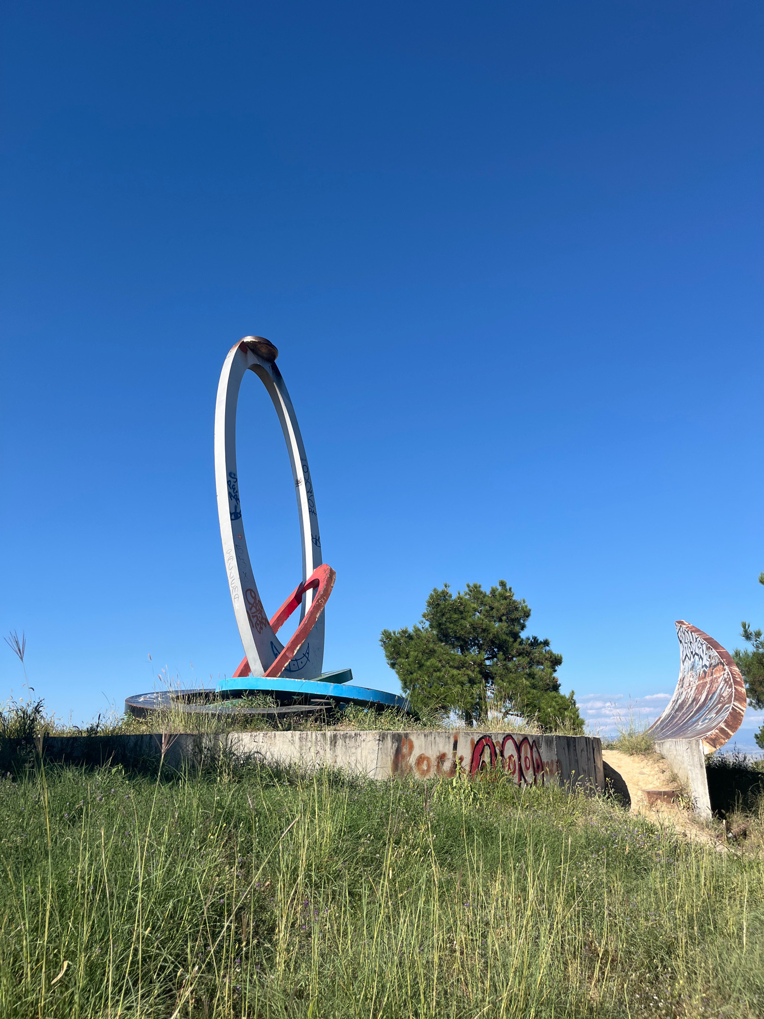 The image depicts a large, abstract metal sculpture with rings, located in an outdoor, grassy area with a clear blue sky overhead. The sculpture appears weathered and is partially covered in graffiti. In the background, there are trees and a distant landscape. The sculpture has a white, large ring, a red inner ring, and a blue base. To the right, there's a curved, rusty metal structure, also with graffiti. The area is overgrown with tall grass and some small weeds. The sculpture is situated on a concrete platform. The scene appears to be in a somewhat neglected or abandoned state. There are no people visible in the image. The location is identified as the Chronicles of Georgia monument in Tbilisi, Georgia, although this specific part of the monument is not prominently featured in typical tourist photos and appears to be a lesser-known or perhaps unfinished section.