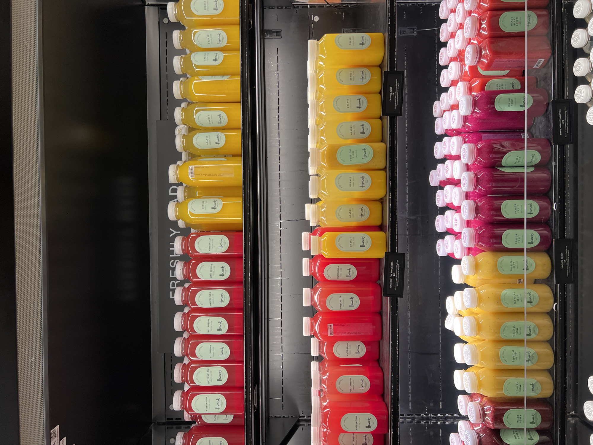The image displays three refrigerated shelves filled with juice bottles, likely inside a shop or deli. The bottles are arranged in neat rows. Each bottle has a label reading "Harrods". The available flavors include watermelon, orange, tropical glow, dragon fruit, and mango berry breeze. The shelves are illuminated by internal lighting. Text on labels indicate prices. The context of the Harrods branding suggests the location may be within Harrods department store in London, UK.