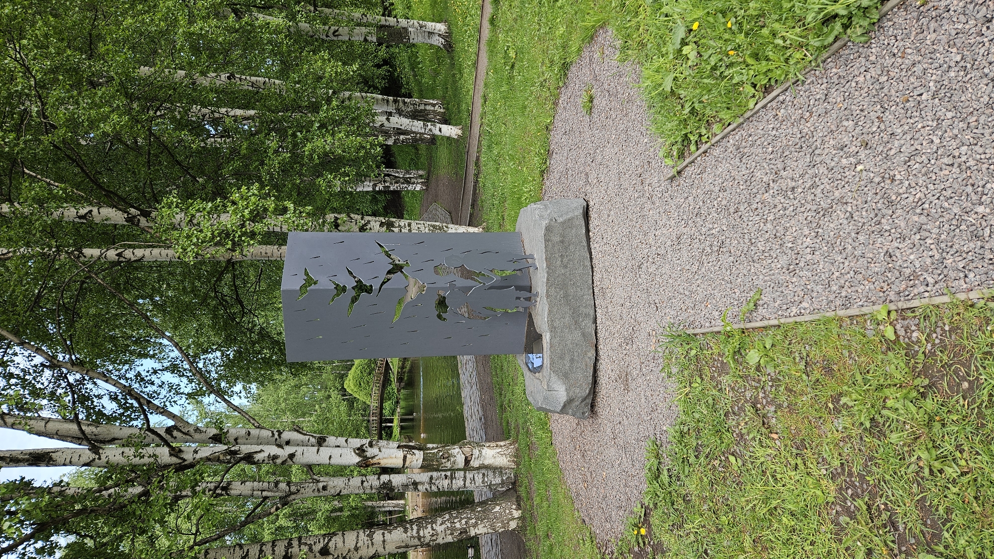 The image shows a gray stone monument along a gravel path in a park, surrounded by birch trees. The monument features cut-out bird silhouettes and a stone base with a water feature. In the background, there's a pond with a bridge. The location is a park, possibly in Unknown, Unknown, given the monument's presence. The environment is green with grass and trees, suggesting a natural or landscaped area. There is no human presence in the picture.
