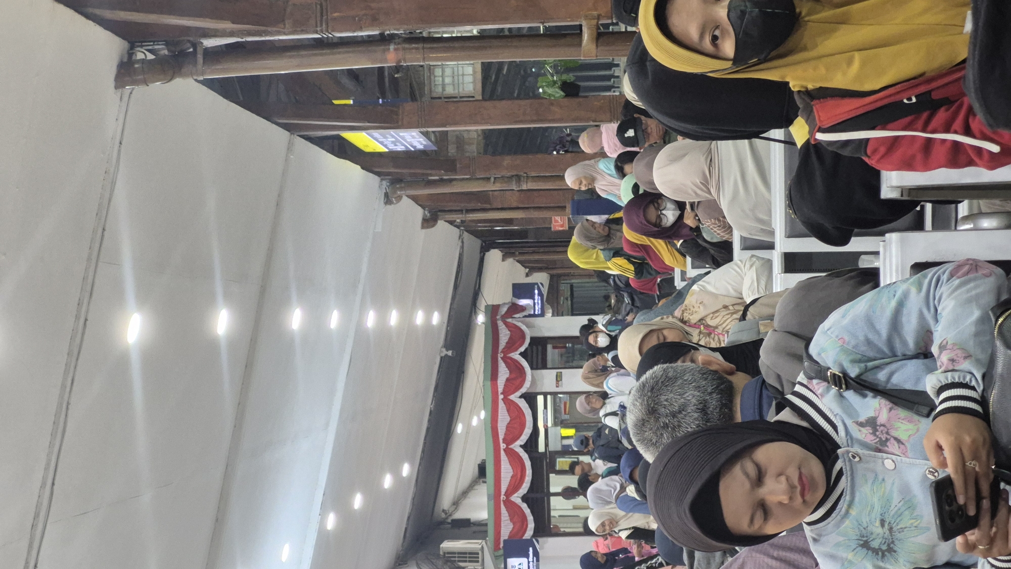 The image depicts a crowded waiting area, likely a train station, with numerous people seated and standing. Many individuals are wearing hijabs, suggesting a predominantly Muslim population. The lighting appears to be artificial, with rows of recessed ceiling lights. Wooden pillars support the roof structure, and there are signs and windows visible in the background, hinting at the station's interior. The atmosphere seems to be one of waiting, with many people looking at their phones or interacting with companions.
While the image itself does not provide explicit information to pinpoint its exact location in Bojonegoro, Indonesia, its characteristics are consistent with a public transportation hub in the region. The presence of Indonesian flags on the facade of a building in the background suggests a connection to Indonesia. The architectural style and the attire of the people are also typical of Southeast Asia. Therefore, it is plausible that this scene is from a train station in Bojonegoro, Indonesia, such as Stasiun Bojonegoro, which serves as a major transportation point in the city. The overall scene conveys a sense of everyday life and travel within an Indonesian urban environment.