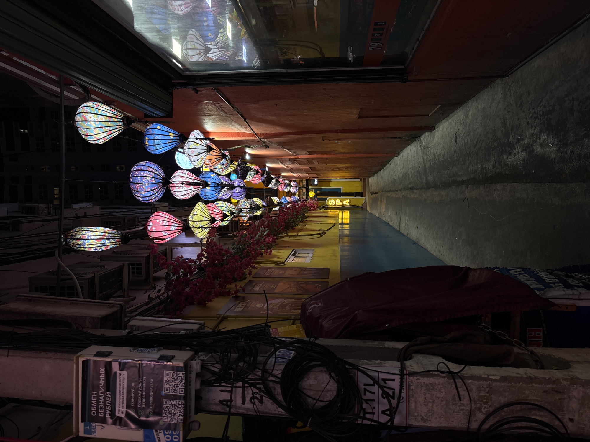 The image depicts a narrow alleyway at night, illuminated by numerous colorful lanterns hanging overhead. The alley is lined with buildings featuring vibrant, textured walls in shades of yellow and red. Electrical wires and conduits are prominently visible along the left side of the alley, attached to a concrete pillar. A sign with Russian text and QR codes is mounted on this pillar. The ground is paved with concrete, showing signs of moisture. On the right side of the alley, a building with a large glass window and red framing is visible, with what appears to be signage or reflections on the glass. The lanterns are varied in shape and pattern, contributing to a festive and somewhat disorienting atmosphere. There are no people visible in the scene. The overall impression is that of a commercial or entertainment district, possibly with a focus on tourism, given the international language on the sign and the decorative lighting. The context of Nha Trang Ward, Vietnam, suggests a Southeast Asian urban environment.