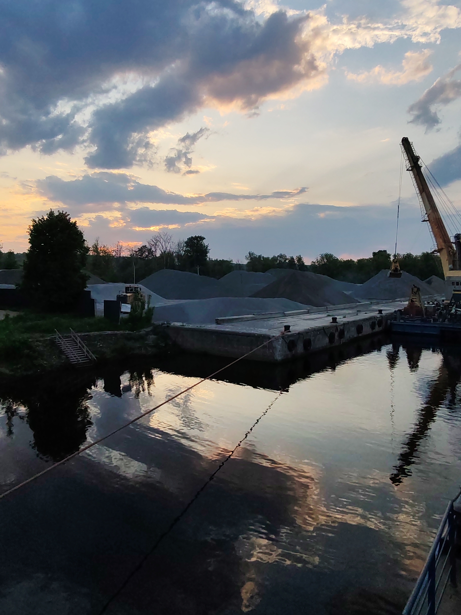 The image depicts a waterfront scene in Yakhroma, Russia. A concrete pier with mooring bollards extends into the water. Behind the pier are several large piles of gravel. A crane is positioned on a barge alongside the pier, with its boom extended over a gravel pile. Trees line the horizon in the background. The sky is filled with clouds, suggesting either sunset or sunrise, reflected in the water. A rope extends from the lower left corner of the image to the pier, likely from the viewpoint of the camera.